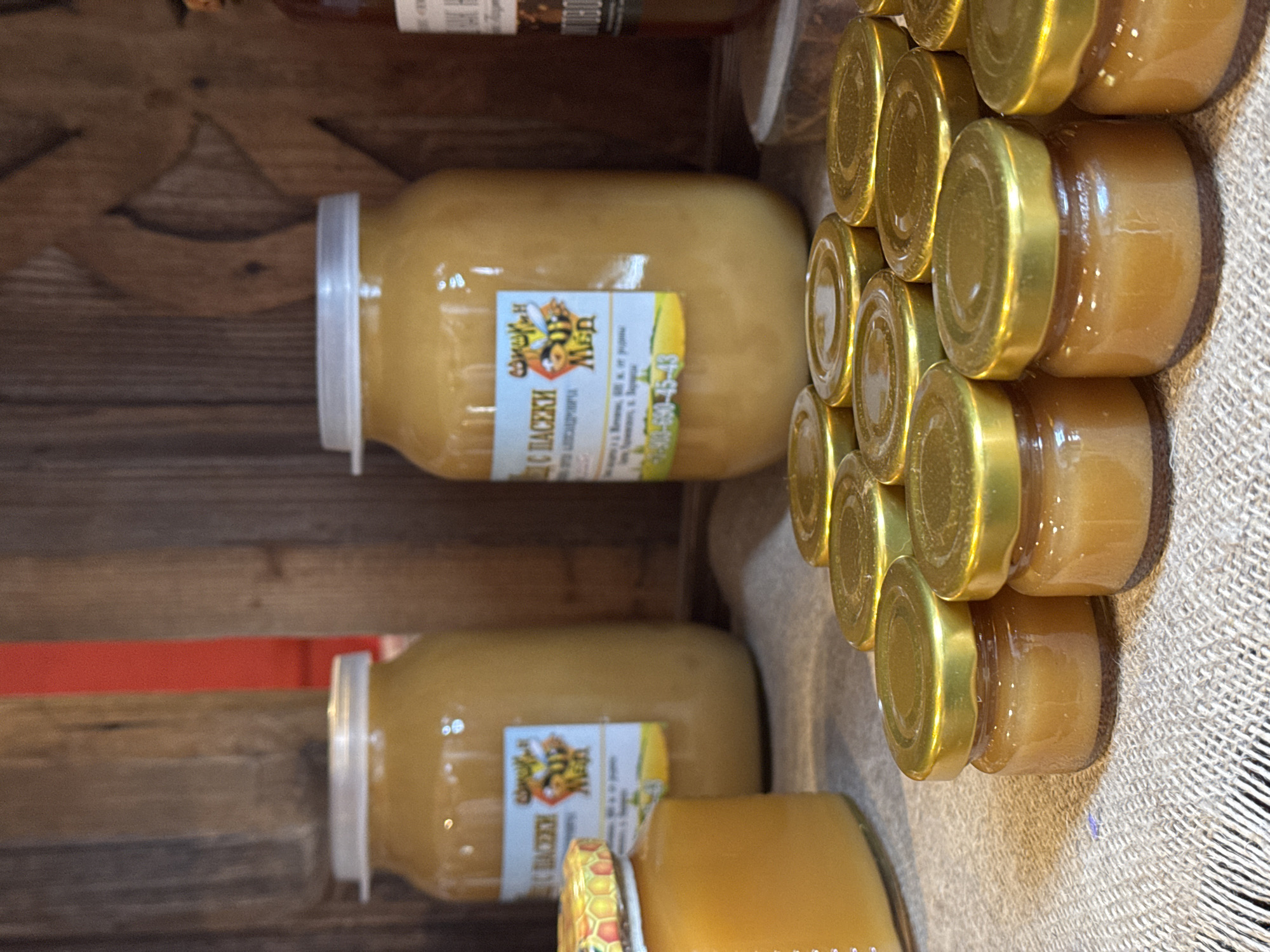 The image shows shelves containing jars of honey. Larger jars with white lids and labels written in Cyrillic are stacked alongside smaller jars with gold lids. The shelves are wooden, with a red support visible. The location, Novo-Nikol'skoe, Russia, is based on the Cyrillic text on the labels.