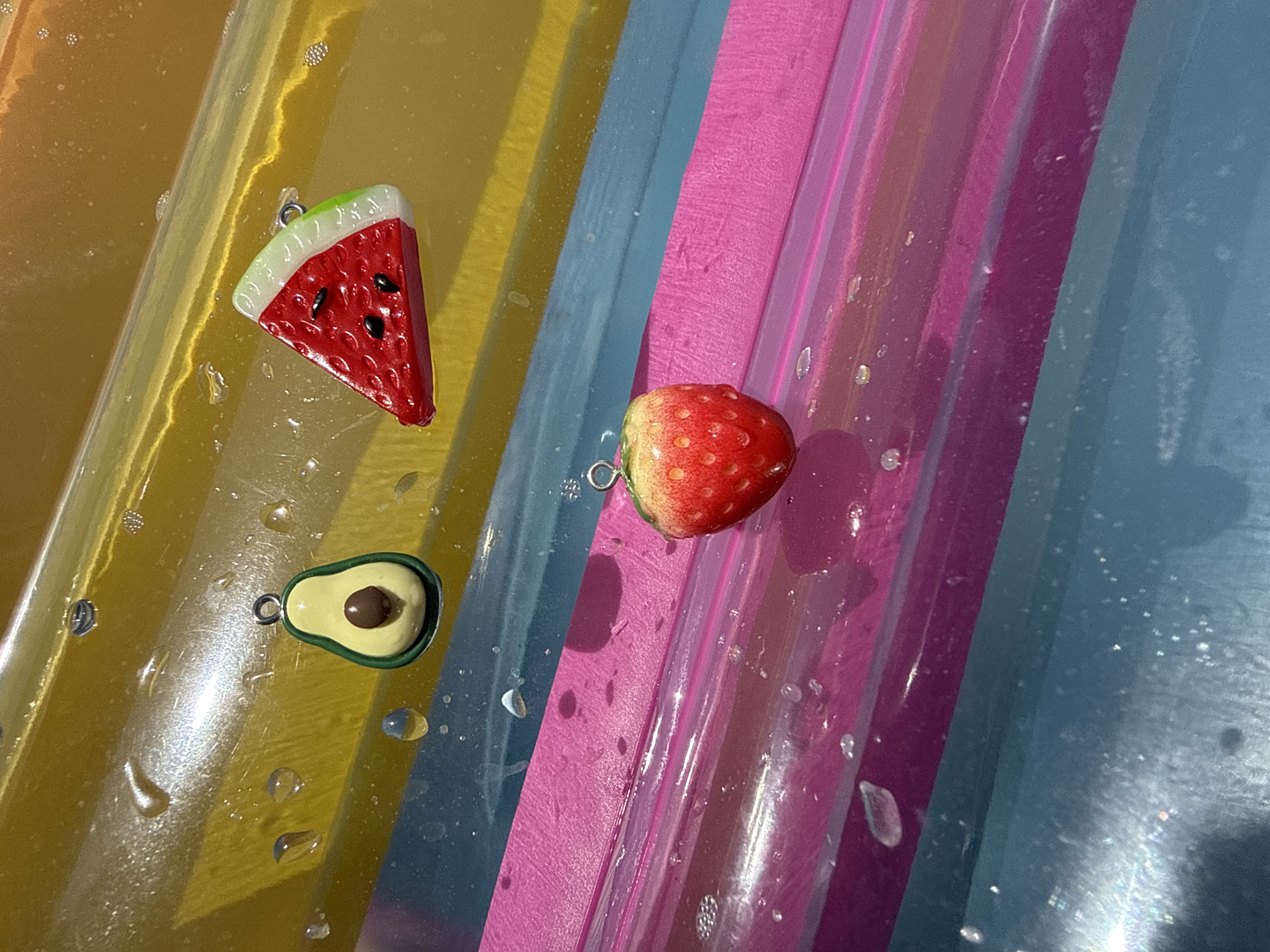 The image displays three small, fruit-shaped charms: an avocado, a slice of watermelon, and a strawberry. These charms are positioned on a surface that appears to be composed of horizontal, colored stripes, likely a plastic inflatable or similar material. Water droplets are visible on the surface, indicating a wet environment. The charms are detailed and appear to be made of a resin or plastic material, each with a small loop for attachment. The avocado charm is green with a brown pit. The watermelon charm is red with black seeds and a green and white rind. The strawberry charm is red with yellow accents and visible seeds. The background consists of alternating bands of yellow, blue, and pink, with some glossy reflections and scattered water droplets. There are no people or indications of a specific location within Gara Oreshets, Bulgaria.