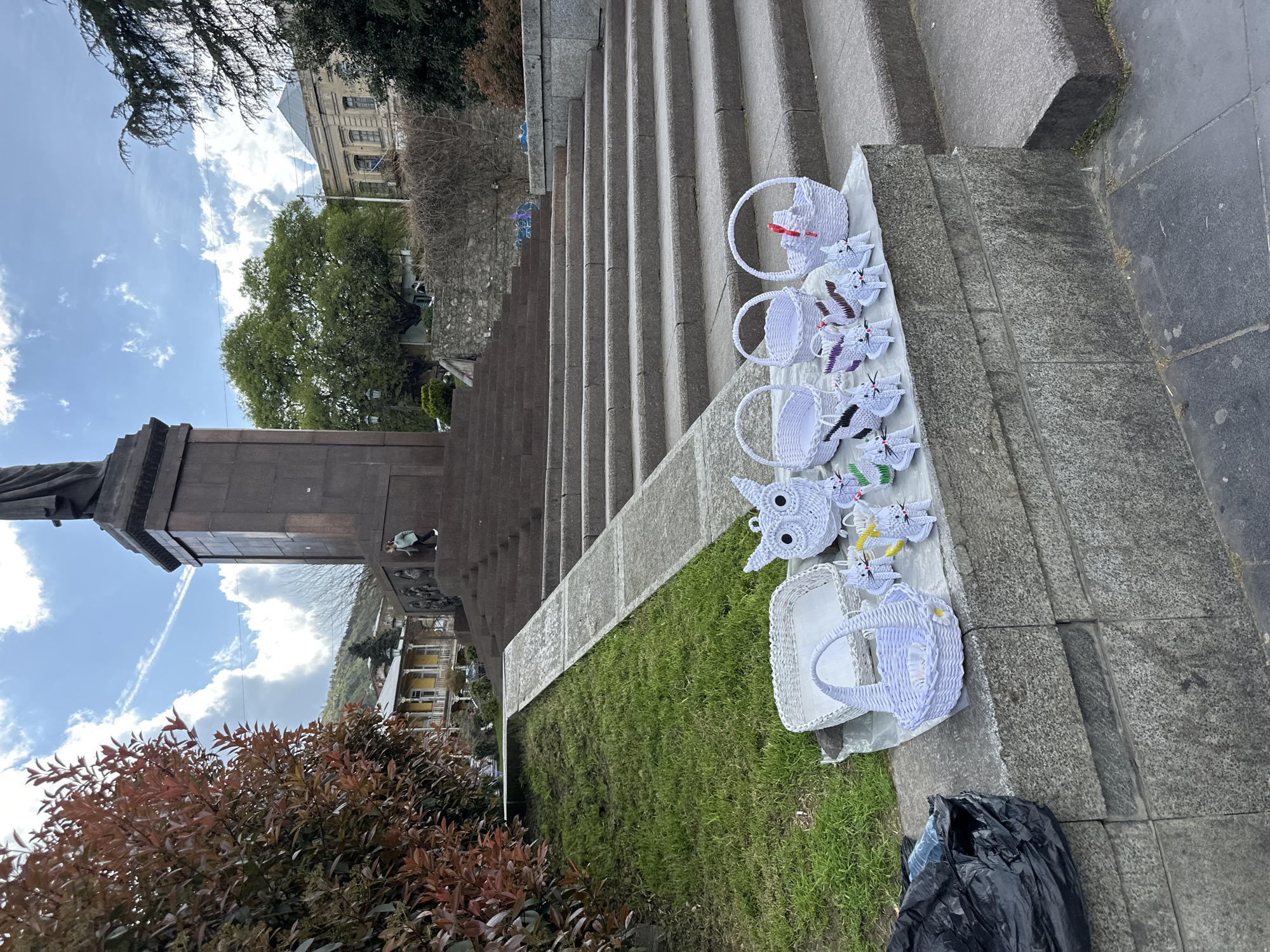 The image shows a vendor selling woven baskets and origami birds on the steps of a monument in Tbilisi, Georgia. The baskets and origami are white, with some color accents. The monument is made of brown stone and is situated at the base of a long staircase. In the background, buildings and trees can be seen. The sky is blue with some clouds. A black plastic bag sits at the bottom right corner of the image, and a bush is in the bottom left. There is no person visible from the chest up, except for a person in the back who is wearing a green shirt and black pants.