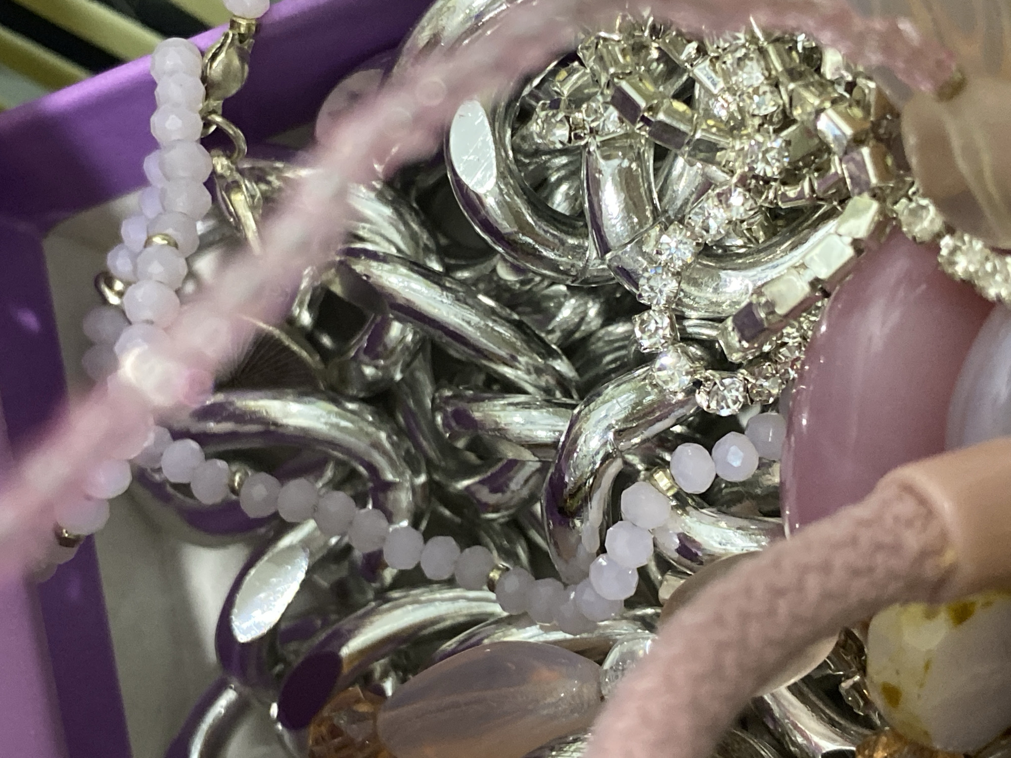 A close-up image shows a jumble of jewelry, including silver-colored chain links, beaded strands (primarily pink or light purple), and pieces with embedded crystals or rhinestones. The jewelry appears to be stored in a container with a purple interior. The lighting is somewhat diffused. There is no indication of a specific location within Chaikovsky, Russia, or any other city. The image focuses exclusively on the jewelry pieces and their arrangement. There are no discernible people, actions, or interactions captured in the frame.
