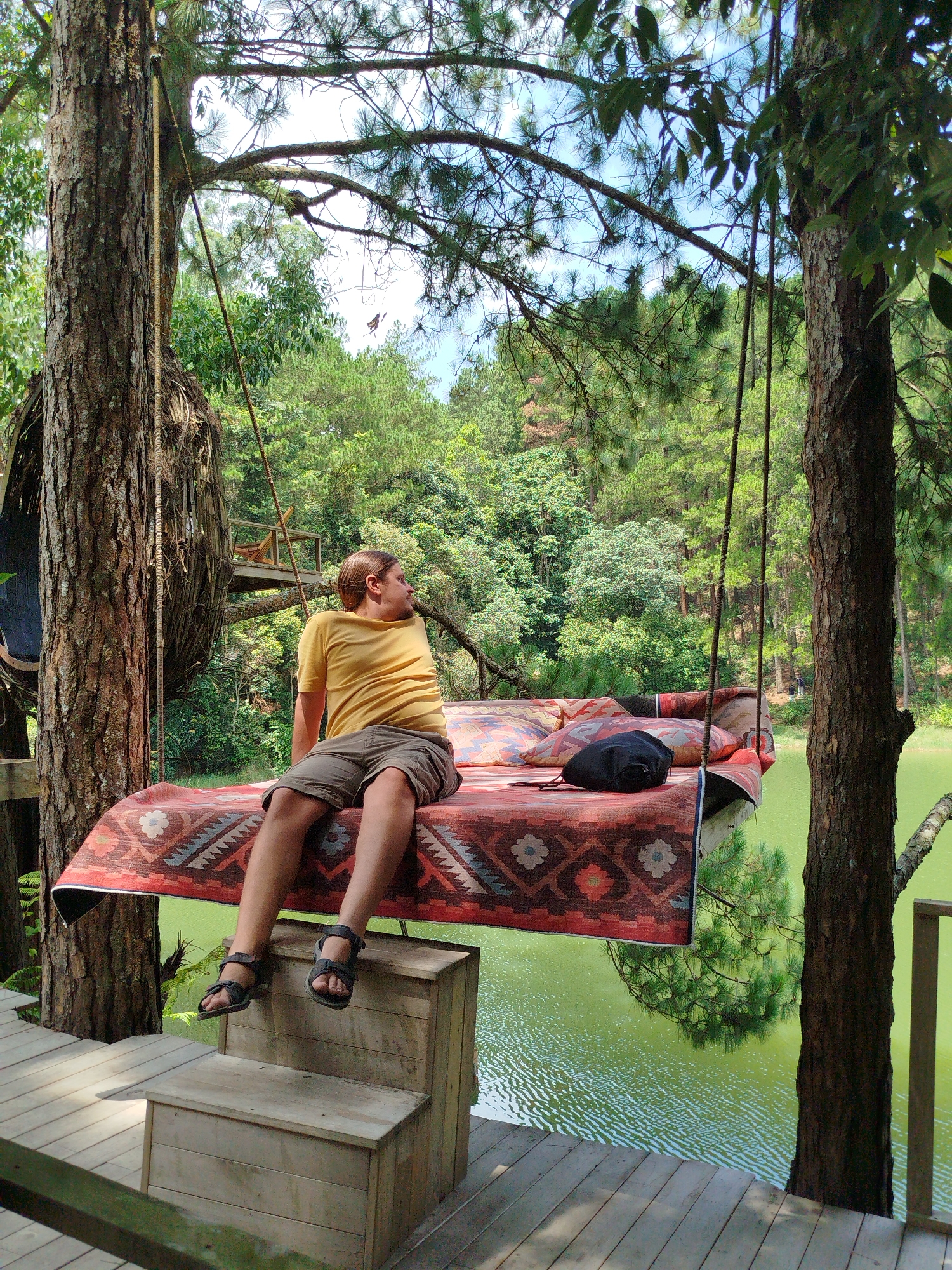 A man in a yellow t-shirt and grey shorts is sitting on a hanging bed made of wood and covered with a patterned rug. The bed is suspended by ropes attached to trees. The man is looking to his right, away from the camera. He is wearing sandals. In the background, there is a body of green water, a forest of pine trees, and a wooden platform. The scene appears to be at Cam Ly - Da Lat, Vietnam.