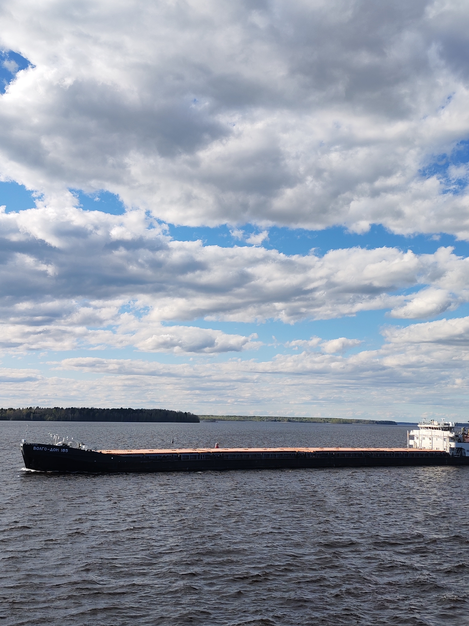 The image shows a freighter ship, labeled "ВОЛГО-ДОН 185" on its bow, navigating a body of water near Rechnaya Sosnovka, Russia. The vessel appears to be carrying cargo. A tree line is visible on the horizon in the distance. The sky is filled with cumulus clouds with patches of blue sky. The water is dark, and the image is taken during daylight hours. No people are visible on deck.