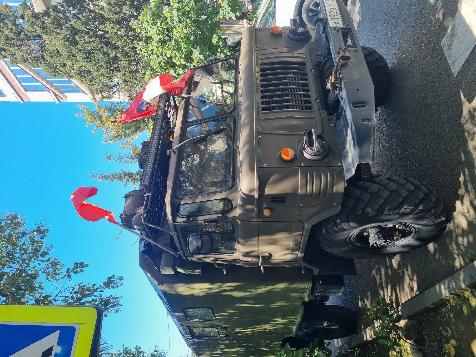 The image shows a GAZ-66, a Soviet/Russian 4x4 all-road (off-road) military truck, parked on what appears to be a street in Sochi, Russia. The vehicle is olive green, and modified with a roof rack and off-road tires. It has a license plate that appears to read "X192BT193". There are red and white flags attached to the vehicle. The sky is blue. Trees and a building are visible in the background. The scene suggests the truck is near a sidewalk or pedestrian crossing.