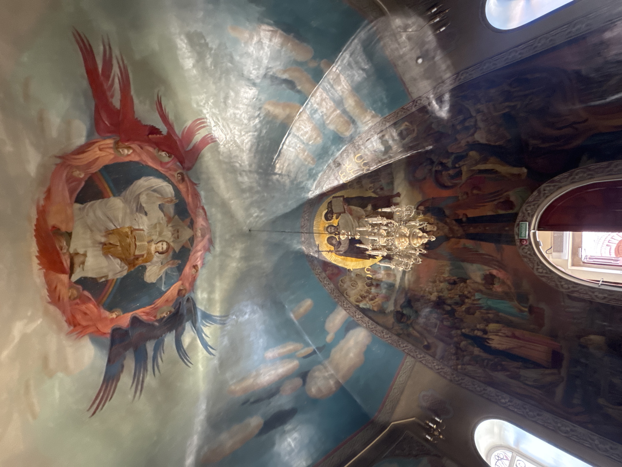 The image shows the interior of a domed ceiling in a place of worship, likely a church or cathedral, adorned with intricate religious murals. The central feature of the ceiling is a large circular fresco depicting a divine figure surrounded by winged beings, possibly angels or seraphim, with vibrant red and blue wings. Below this, the ceiling continues with a fresco of a procession of figures, with a chandelier hanging in the foreground, illuminated by light filtering through arched windows on either side. The walls are also covered with large-scale murals, appearing to depict biblical scenes and religious figures. The overall impression is one of grandeur and spiritual devotion, with the artwork dominating the space and creating a sacred atmosphere. The location is described as Unknown, Unknown.
