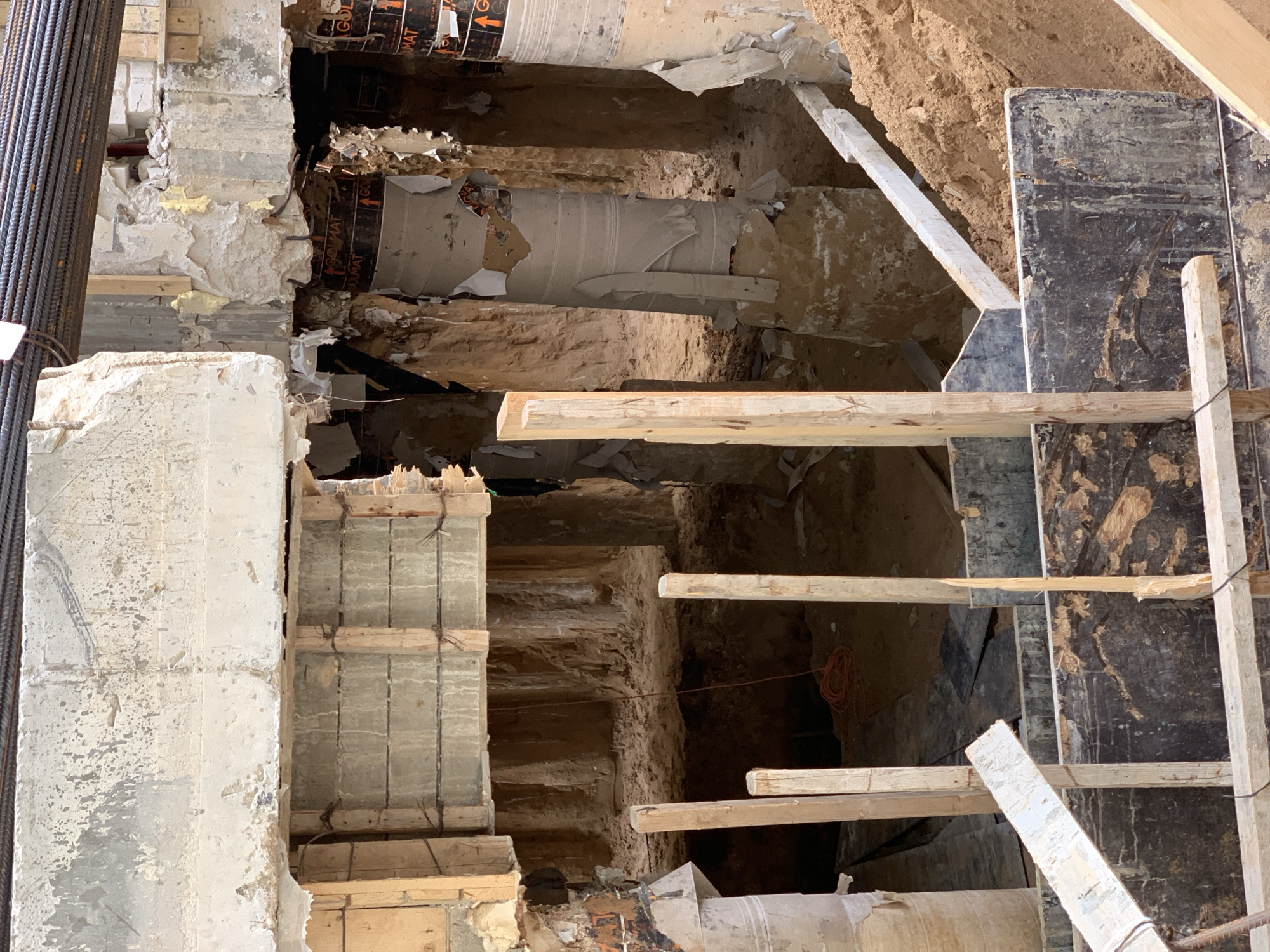 The image depicts a construction site, potentially in Tel-Aviv, Israel, though specific location cannot be confirmed solely from visual elements. The scene features exposed building foundations, revealing various layers of materials including concrete, brick, and insulation. There are sections of damaged or removed walls, exposing what appear to be underground pipes or conduits wrapped in protective material. Wooden supports and scaffolding are present, suggesting ongoing construction or demolition work. The environment consists of excavated earth and debris, indicating active site modification. No people are directly visible in the frame. The focus is on the structural components and the process of excavation or rebuilding.