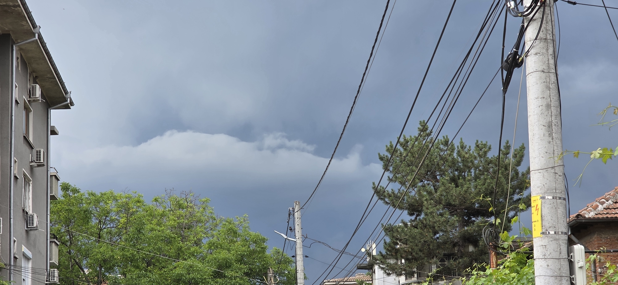 This outdoor, eye-level shot captures a street scene in Plovdiv, Bulgaria, under an overcast and potentially stormy sky. The lighting suggests daytime, but the heavy, dark grey and white clouds dominate the upper half of the frame, hinting at impending rain or an approaching storm. A distinct horizontal cloud band stretches across the mid-sky.
On the left, a grey, multi-story residential building is partially visible, featuring multiple windows and several external air conditioning units mounted on its facade. A roofline with a gutter system is also discernible.
Towards the center and bottom of the image, lush green foliage from two types of trees is present: one with lighter, deciduous leaves and another with darker, needle-like foliage, likely a pine. These trees add natural contrast to the urban elements.
The right side of the frame is dominated by a sturdy, concrete utility pole. This pole is heavily laden with numerous black electrical and communication cables that crisscross the cloudy sky. A white, rectangular utility box is affixed near the base of the pole. Vibrant green vines, possibly grapevines, with lighter green leaves, grow around the pole and extend into the foreground.
A yellow warning sticker is prominently displayed on the utility pole. It features a black lightning bolt symbol and Cyrillic text, reading "ОПАСНОСТ" (Danger) and "ВИСОКО НАПРЕЖЕНИЕ" (High Voltage), followed by voltage information like "380 / 220 V" and potentially an emergency phone number. In the background on the far right, part of a building with a reddish-brown tiled roof and a brick wall can be seen. The overall scene depicts a typical urban infrastructure setting under dramatic weather conditions, with no human activity taking place.