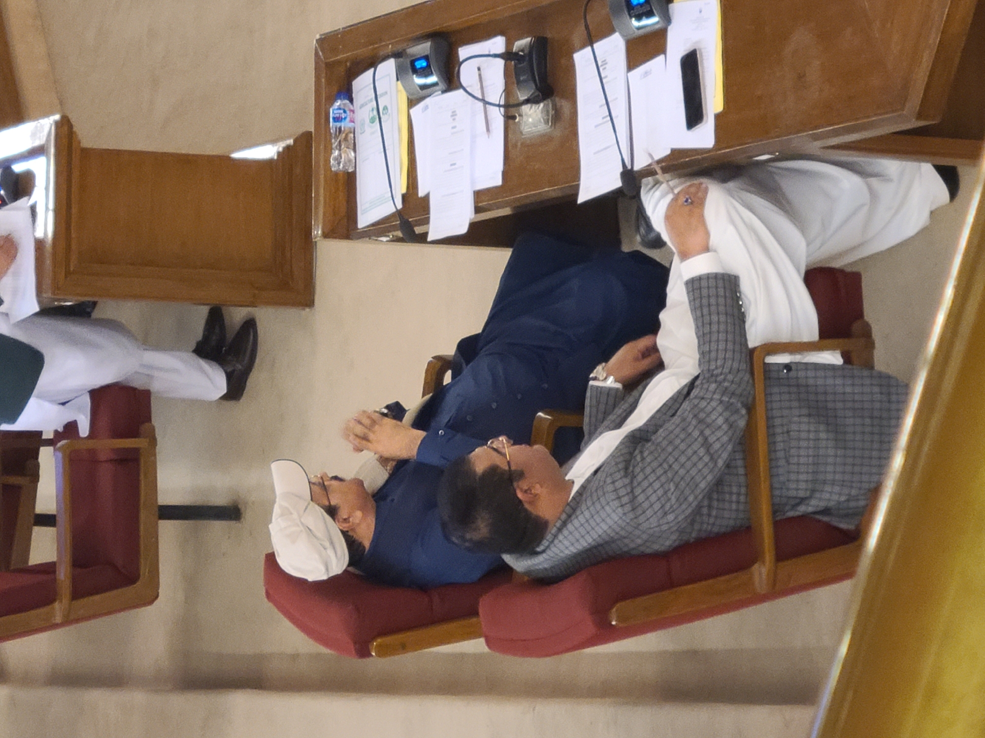 Two men are sitting in red, cushioned chairs facing a wooden desk that holds two black telephones and a bottle of water. Both men are wearing suits and glasses. The man on the left is wearing a white cap. The man on the right is wearing a gray plaid suit jacket. Another man is partially visible in a chair on the left. The scene is Quetta City Tehsil, Pakistan.