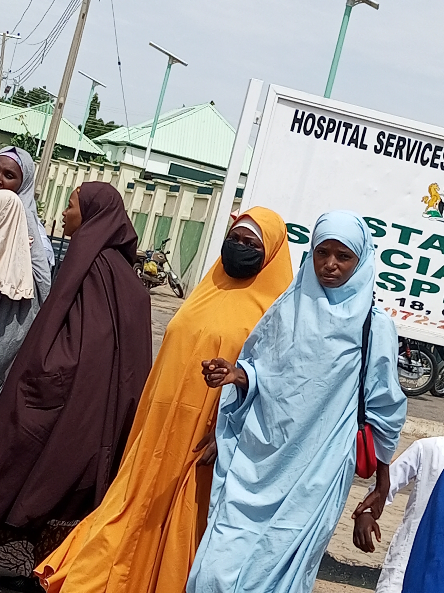 In Arawa, Nigeria, on a bright day, four women are walking past a building with a sign that reads "HOSPITAL SERVICES". The woman on the far right wears a light blue hijab and a light blue garment, with a red purse slung over her shoulder. To her left, a woman in an orange hijab and mask is visible. Next to her, a woman in a dark brown hijab and garment walks with her head turned slightly. The woman on the far left is mostly obscured, wearing a light-colored hijab. The background shows a row of buildings with green roofs and decorative fencing. A motorcycle is parked on the side of the road. Overhead, electrical wires are strung between utility poles, one of which has a solar panel attached. The ground is paved, and a curb is visible.