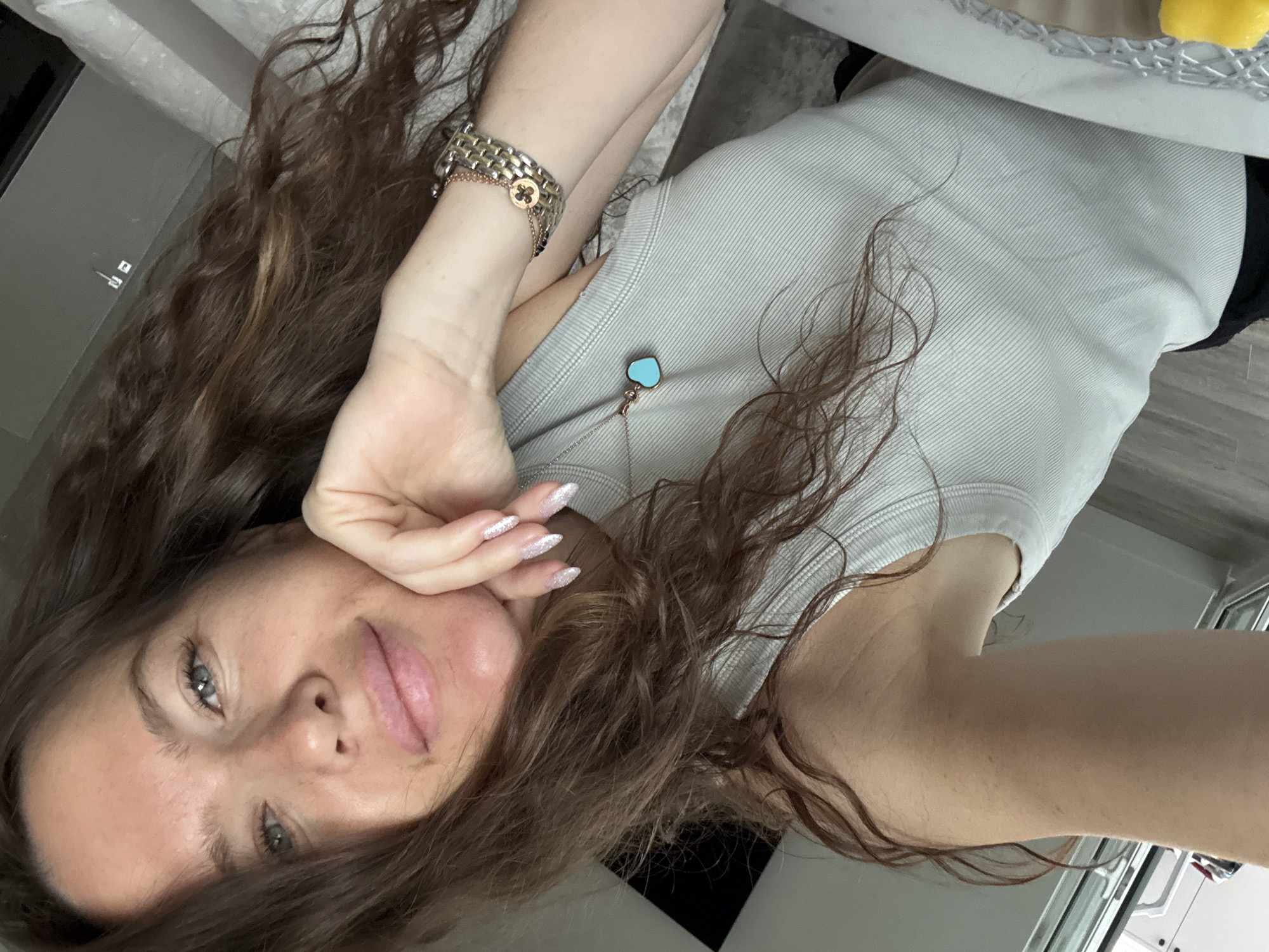 A young woman with long, wavy brown hair is taking a selfie indoors. She is wearing a light grey ribbed tank top and a silver bracelet. A turquoise heart-shaped pendant necklace is visible. Her left hand is resting on her chin, and her nails are painted with silver glitter polish. The background is blurred, but appears to be a room with a bed and a window. The scene is likely in a residential setting, possibly an apartment or hotel room, and the woman is indoors. The location is not specified in the image.