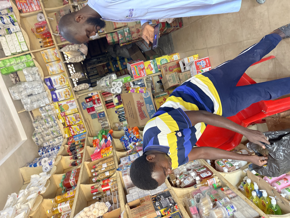 Inside a retail shop in Samaru, Nigeria, two adult males are present amidst shelves heavily stocked with various consumer goods. In the foreground, a male with short dark hair, wearing a horizontally striped polo shirt (blue, yellow, white) and dark blue trousers, is bent over a red plastic chair, actively reaching into a black plastic bag. To the right and slightly behind him, a bald, bearded male, dressed in a light-colored traditional garment, stands upright, looking down at and operating a smartphone held in his left hand. The shop walls are lined with multiple wooden shelves displaying an assortment of products including cooking oils, cereals (Kellogg's Corn Flakes, Golden Morn), packaged snacks (Munch Cheesy Stix, Indomie noodles), personal care items, cleaning supplies, and bulk goods like toilet paper and flour/sugar bags. Bunches of garlic hang from the ceiling in mesh bags. The floor is visible as a light-colored surface, and the overall environment is well-lit.