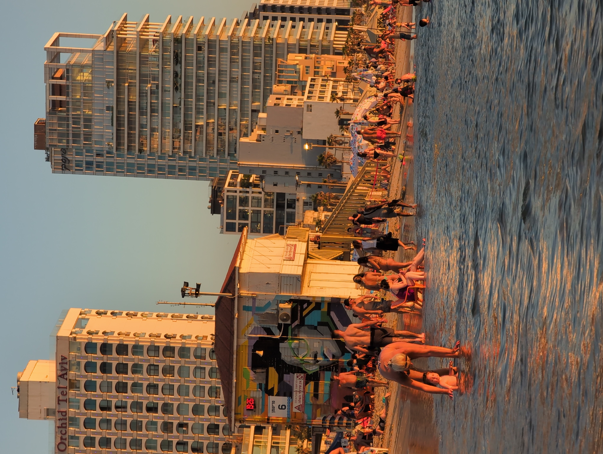 This is a horizontal, eye-level, medium shot photograph of a beach in Tel Aviv, Israel, during a sunset. The golden hour light casts a warm glow on the scene, illuminating the water, sand, and buildings. In the foreground, a woman is bending over to play with a baby in the shallow water. The beach is populated with numerous people, some sunbathing, others walking or standing. Behind the beach, a white building with colorful graffiti on its side stands out, possibly a lifeguard station or beach concession. In the background, modern high-rise buildings with contemporary architecture dominate the skyline, reflecting the sunset. The overall atmosphere is one of a busy, yet relaxed, urban beach scene.