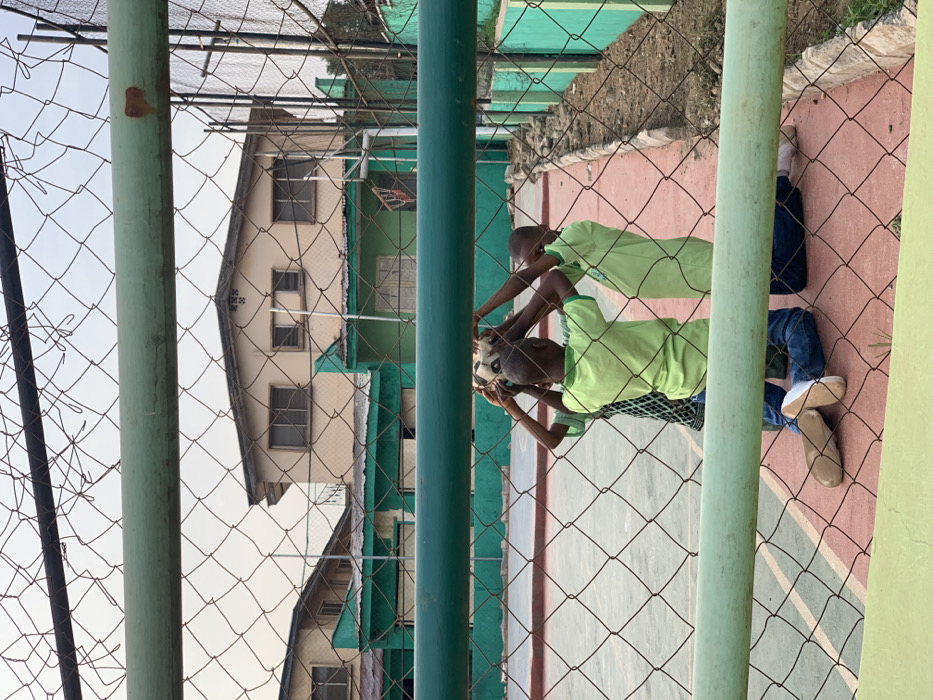 Three young boys are playing with a soccer ball in a basketball court in Akure, Nigeria. They are kneeling and holding the ball above their heads. The court is surrounded by a chain-link fence and green walls. The fence is visible in the foreground, and the court surface is red and lined with white. Two of the boys are wearing green shirts, while the third is wearing a light blue shirt. The image is taken from a slightly elevated position behind the fence. The boys are interacting and laughing, appearing to be having fun while playing. The scene is located in a residential area, with a building in the background. The building is a two-story structure with a flat roof and several windows. There are also some trees and bushes around the court. The sky is clear and blue. The image is likely taken in the daytime, and the lighting is good. The image is in focus and sharp.