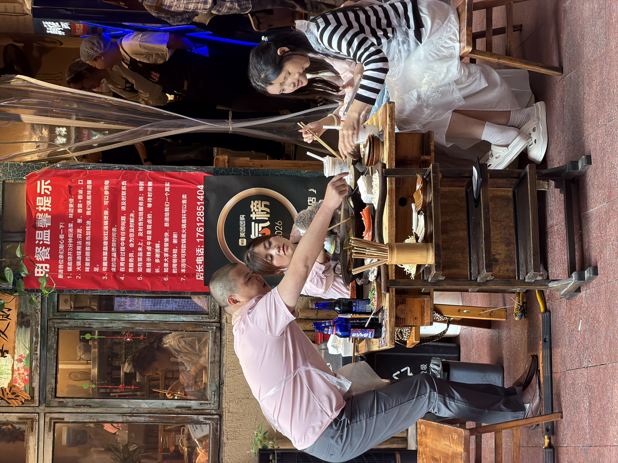 In the Yuzhong District of China, an outdoor restaurant scene unfolds. A man in a pink shirt and grey pants stands over a table, gesturing with chopsticks towards a meal being prepared by two young women. One woman, wearing a black and white striped top, is focused on her food, while the other, in a pink top, looks towards the man. The table is laden with dishes, including what appears to be hot pot ingredients, chopsticks, and drinks. A red sign with Chinese characters hangs prominently in the background, offering dining instructions and a customer service phone number. The setting suggests a busy eatery, with other patrons visible in the blurred background and a street scene visible through a window. The overall atmosphere is one of casual dining and interaction.
