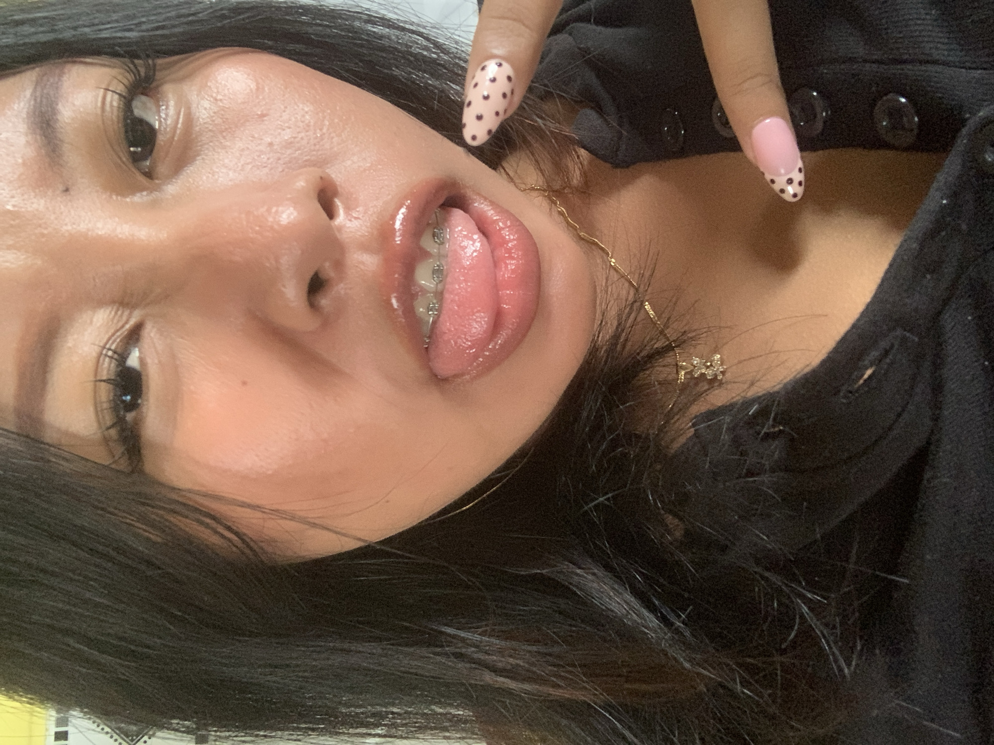 The image is a close-up selfie of a young woman with dark hair and braces. She is wearing a black top with a low neckline and a gold necklace. Her tongue is sticking out, and her nails are painted pink with black polka dots. The image appears to have been taken indoors, as suggested by the lighting and the background, which is out of focus and shows a wall with a patterned border. There is no indication of the specific location within Jajag, Indonesia.
