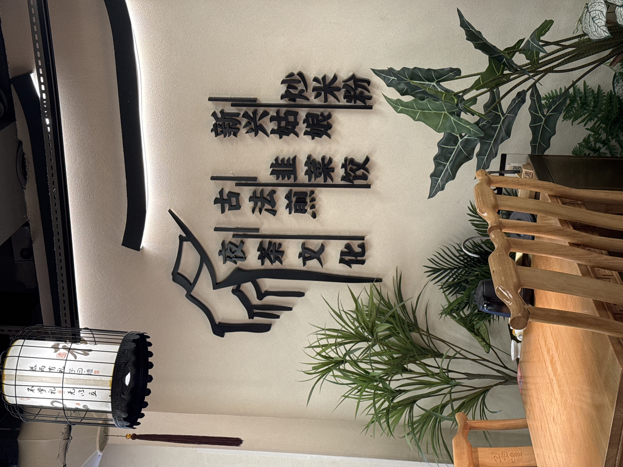 The image shows a wall in Yuexiu District, China, decorated with Chinese calligraphy and plants. A black, decorative lantern with Chinese writing hangs from the ceiling. The calligraphy on the wall is also in Chinese and appears to be a decorative piece. Two different types of plants are visible on the right side of the wall. A wooden chair is partially visible on the right side of the image, near the plants.