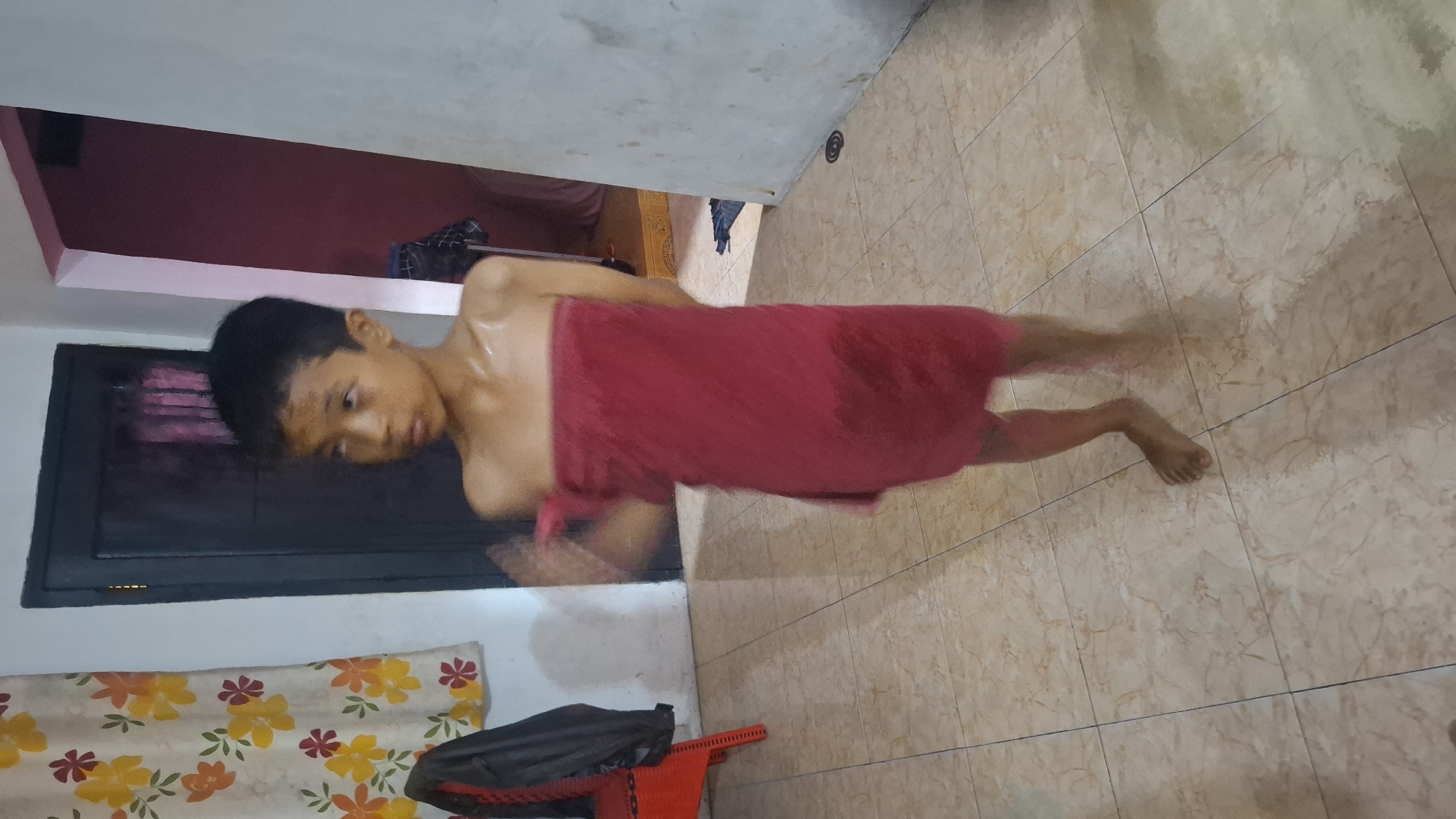 In a room with tiled flooring, a young boy stands wrapped in a red towel. He is looking to his right with a slightly blurred face and body, indicating movement. His shoulders are bare, and the towel covers him from just below his chest down to his knees. The background features a dark-colored door with what appears to be a curtain behind it, and a wall painted pink and white. To the boy's left, a red chair with a black jacket draped over it is visible next to a floral-patterned curtain. The overall lighting suggests an indoor setting. The location is identified as Cibinong Hilir, Indonesia.