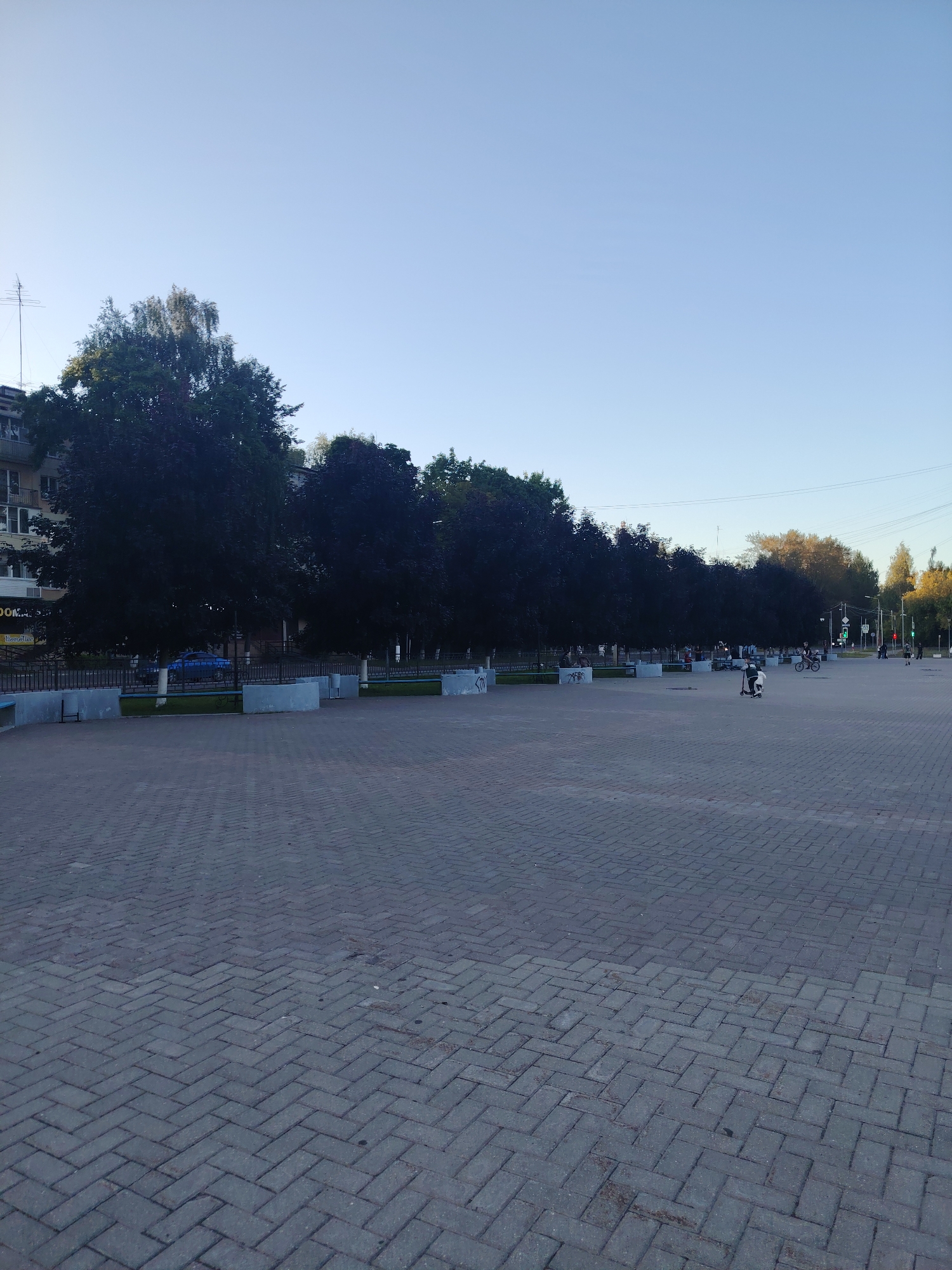 The image depicts a wide, paved plaza in the city of Konakovo, Russia. The plaza is covered with interlocking grey bricks laid in a herringbone pattern. In the background, a line of dark green trees forms a dense canopy. Beyond the trees, a building with multiple balconies and windows is visible, along with some power lines and a radio antenna on the left side. The sky is a clear, pale blue, suggesting it is either early morning or late afternoon.
Several people are present in the plaza, primarily in the middle and right sections. On the right, a person is riding a bicycle and another is on a scooter. Further in the distance, a group of people are gathered, some appearing to be sitting on benches or low concrete structures. These structures are also visible along the edge of the treeline, appearing to be part of a park or a landscaped area. A blue car is parked behind a black metal fence in front of the trees on the left side of the image. Graffiti tags are visible on the concrete bases of the seating areas. The overall atmosphere appears to be calm and leisurely, with people enjoying the open space and the presence of trees.