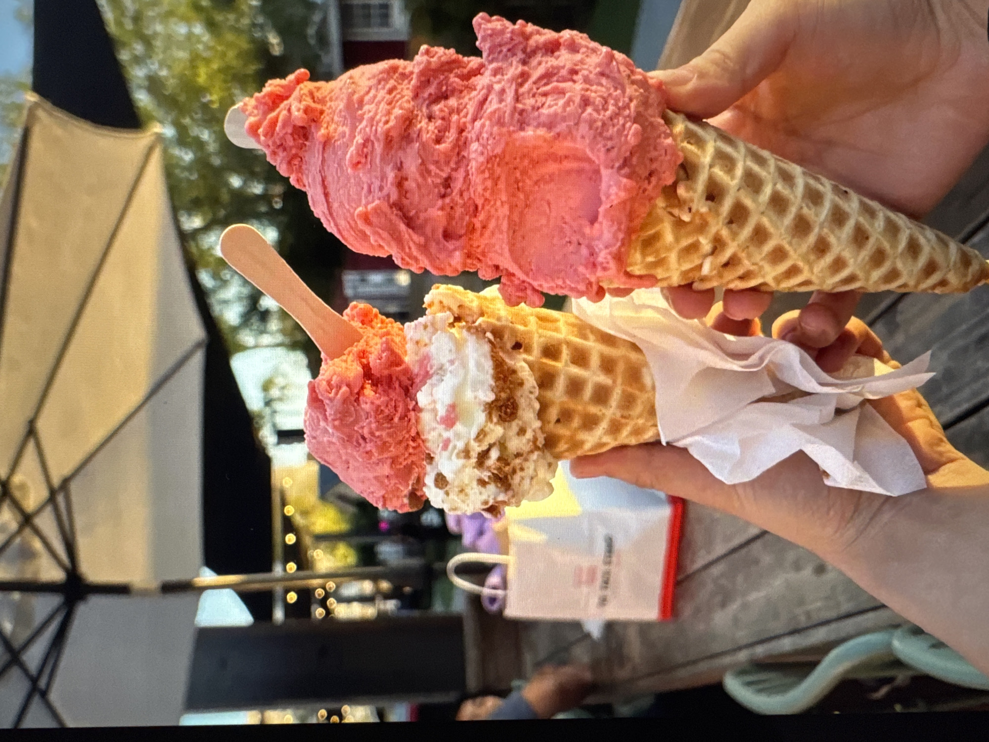 Two hands hold waffle cones filled with scoops of pink ice cream. The ice cream on the left is topped with white whipped cream and crumbled cookies. A wooden spoon is partially submerged in the ice cream on the left. In the background, a white umbrella provides shade over a wooden table. A white paper bag with red accents sits on the table, bearing text that reads "TO TALLINN STORY." The scene is outdoors, with trees and a building visible in the distance. The lighting suggests it is late afternoon or early evening.