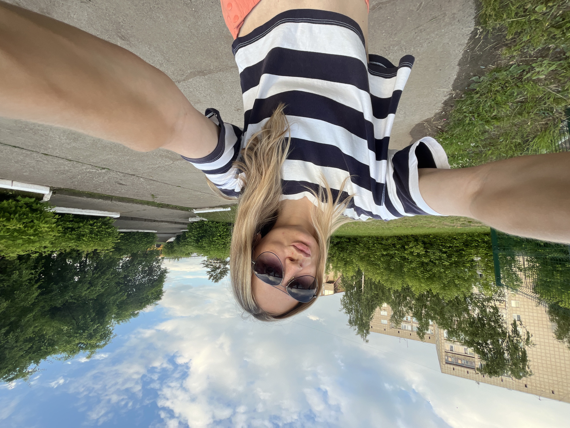 In Chaikovsky, Russia, a young Caucasian woman with blonde hair, wearing a striped t-shirt and sunglasses, is taking a selfie outdoors. She is making a kissing face and is standing on a concrete path. Behind her, there is a green fence, lush green trees, and a multi-story building. The sky above is blue with scattered clouds. The image appears to be taken during the daytime.