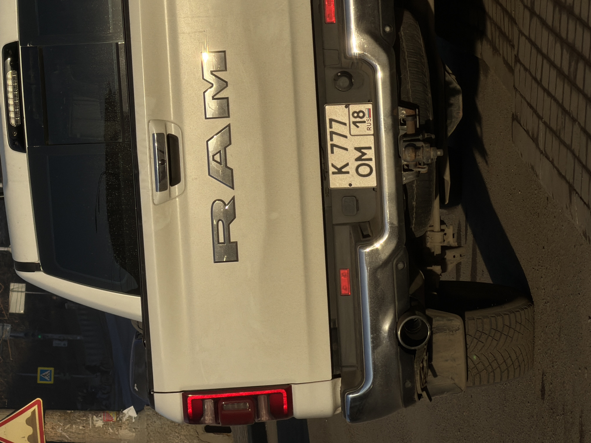 The image depicts the rear of a white RAM pickup truck. The license plate reads "K 777 OM 18" which indicates that this is a Russian vehicle from the region of Udmurtia. Udmurtia's capital city is Izhevsk. The photograph appears to be taken at night or during low-light conditions. The truck is parked near a road and sidewalk. Part of a road sign and a street are visible. The environment appears to be urban.