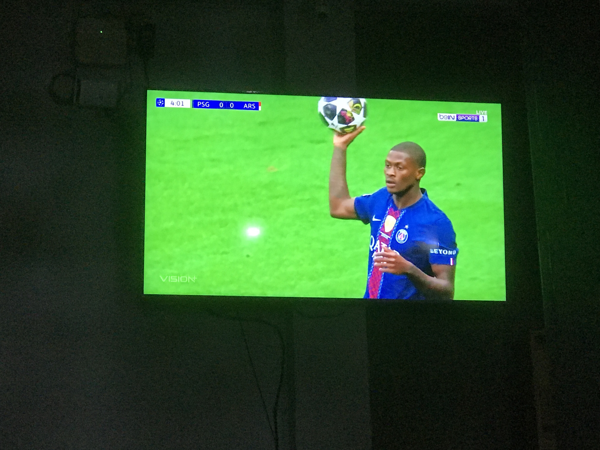 The image shows a television screen displaying a live broadcast of a soccer game. The score is PSG 0, ARS 0, with the game clock at 4:01. A soccer player is shown on the screen, holding a soccer ball above his head. The broadcast is on beIN SPORTS 1. The location is likely a home in Lhokseumawe, Indonesia, as it is inside a room. The environment is dimly lit, focusing the viewer's attention on the screen.