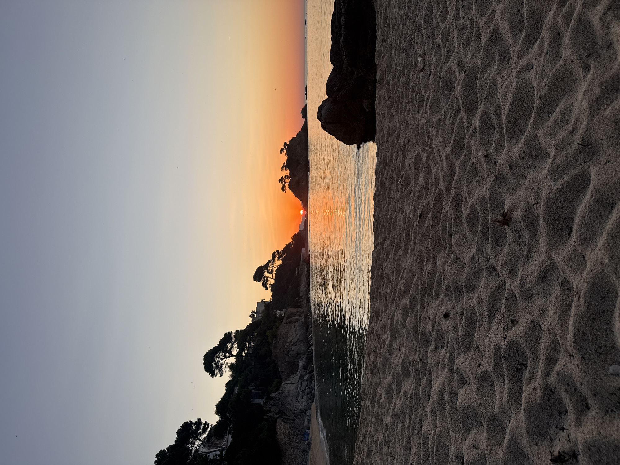 A scenic view of a beach at sunrise is captured in Castell d'Aro, Platja d'Aro i s'Agaró, Spain. The foreground features textured sand with ripples, leading the eye towards the tranquil sea. In the middle ground, a dark, large rock juts out of the water to the right, while the left side shows the calm water gently lapping the shore of a cove. The background is dominated by a rocky, tree-covered coastline with silhouetted pine trees and some buildings nestled amongst the foliage. To the right of the coastline, small islands covered in trees rise from the sea, with the sun just beginning to crest over them, casting a warm orange and yellow glow across the sky and reflecting on the water. The sky transitions from a pale blue at the top to soft oranges and yellows near the horizon, indicating early morning. There are no people or animals visible in the scene. The overall mood is serene and natural.