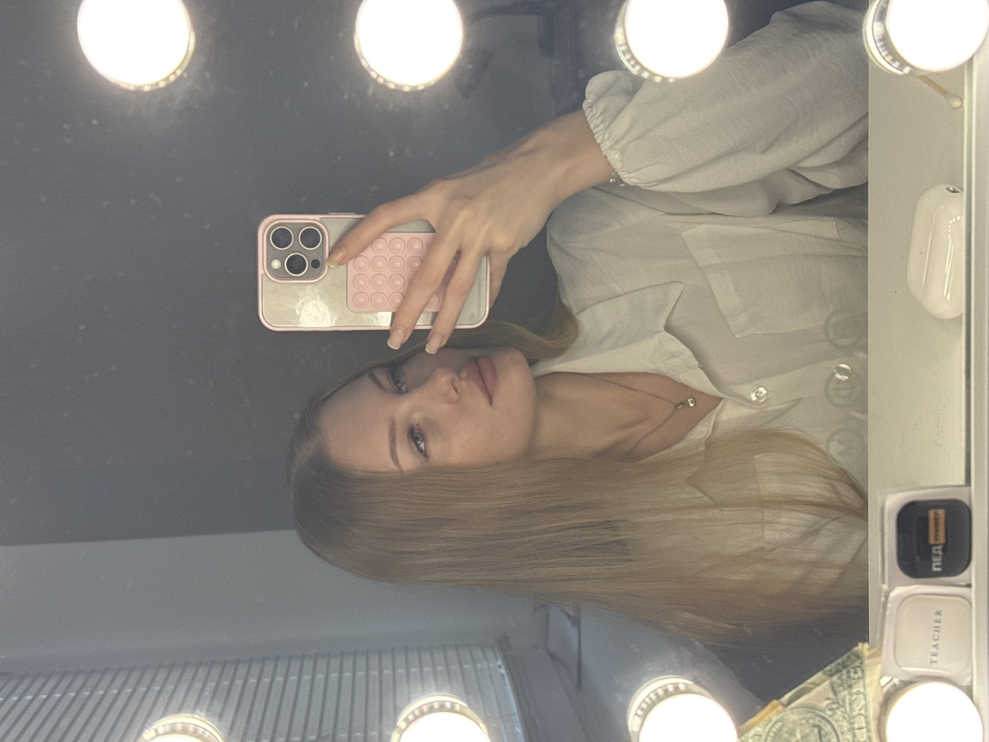 The image shows a selfie taken by a young woman with blonde hair, looking at herself in a mirror. She is wearing a light-colored collared shirt. The mirror appears to be a vanity mirror with a ring of lights around it, typical for makeup application. In the foreground, on a surface below the mirror, are several objects including a phone case with a pink pop socket, a small white container with the word "TEACHER" printed on it, a black case with Cyrillic writing which appears to be "ПЕД," and a white earbud charging case. The background is a wall with some blinds visible on the left side of the image. The overall setting suggests a personal space, possibly a dressing room or bathroom, in a residential or studio apartment. There is no indication of the city of Perm, Russia.
