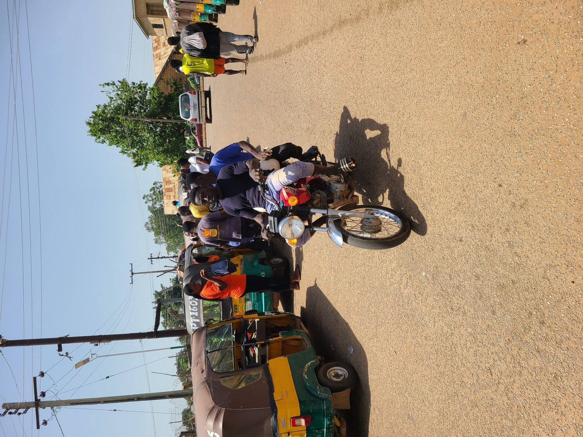 In Bukuru, Nigeria, under a clear sky, a street scene unfolds. A motorcycle, painted red and silver, carries two young men. The driver, wearing a dark t-shirt and striped pants, is focused ahead. Behind him, a second man in a blue t-shirt looks towards the viewer. They are on a paved road, with the rough texture of the asphalt visible. To the left, two yellow and green auto-rickshaws, typical of Nigerian transport, are parked. One has its door open, revealing an interior and what appears to be a cracked windshield. A woman in an orange t-shirt stands by this auto-rickshaw, holding a blue bag. Further down the road, a line of more auto-rickshaws stretches into the distance. On the right side of the road, several people are walking or standing. One man in a bright yellow t-shirt and orange shorts is prominent, as is another man in a grey hoodie and pants with a white bag slung over his shoulder. The background is dotted with buildings, some with corrugated iron roofs, and lush green trees, indicating a warm climate. Electrical poles with tangled wires crisscross the sky, a common sight in urban African landscapes. The overall impression is of a busy, everyday moment in a Nigerian city.