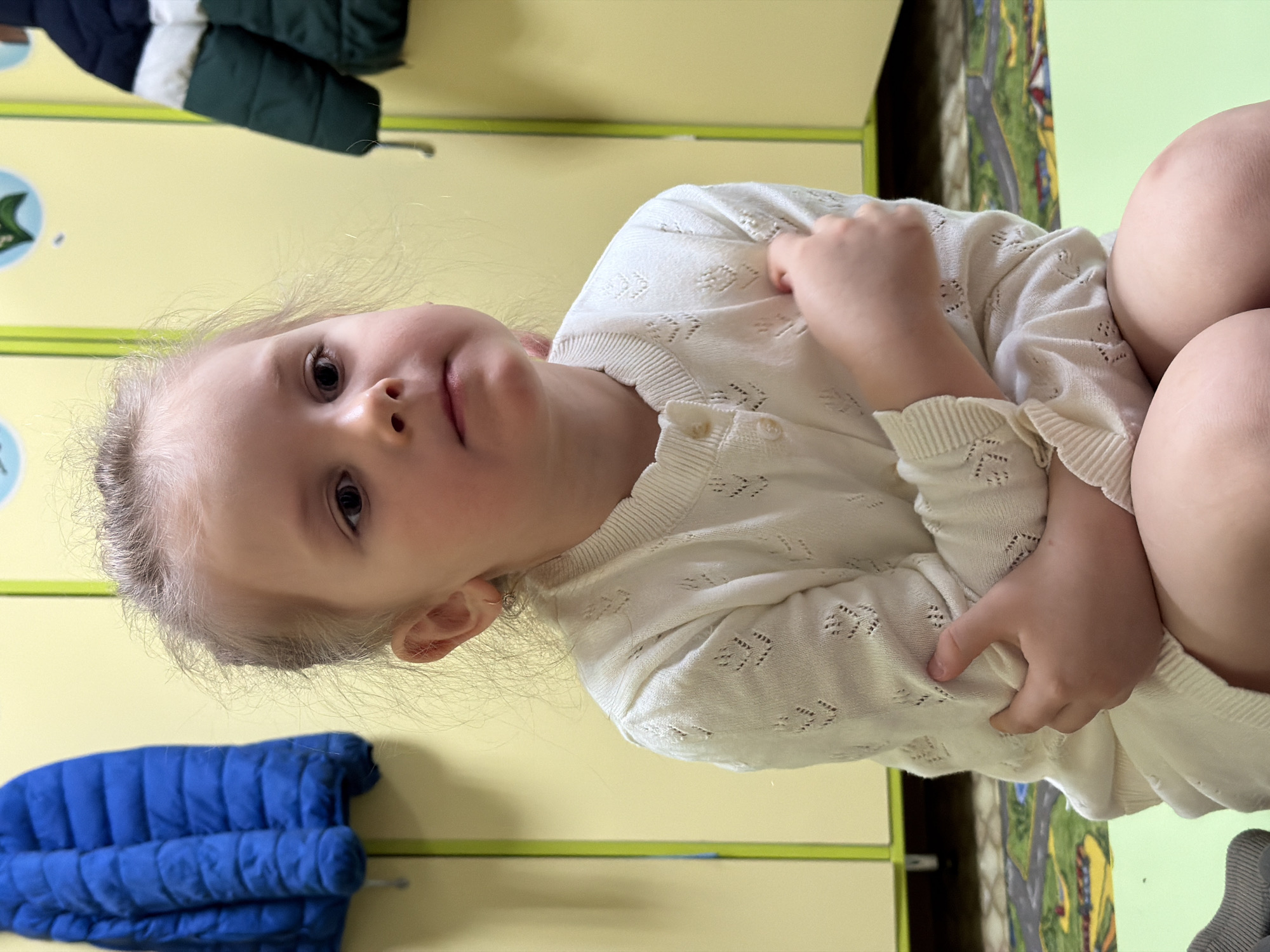 A young child, likely a toddler or preschooler, is depicted sitting and looking directly at the camera in what appears to be an indoor setting in Chaikovsky, Russia. The child has light-colored hair, fair skin, and dark eyes, with a neutral and somewhat curious expression. They are wearing a cream or off-white knitted cardigan featuring an open-work patterned texture and ribbed cuffs. A small, simple hoop earring is visible in the child's right ear.
The background consists primarily of light yellow lockers or cupboards with green trim, suggesting a children's environment such as a daycare, preschool, or playroom. To the child's left (viewer's right), a colorful rug with a road or landscape pattern is visible on the floor. On the far left of the image, a dark blue puffy jacket hangs on a hook, and above it, a dark green and white jacket is also visible, indicating storage for outerwear. The lighting is soft and consistent with an indoor setting. The child's posture is relaxed, with arms possibly crossed or one hand resting on the other arm or leg. No text is visible in the image.