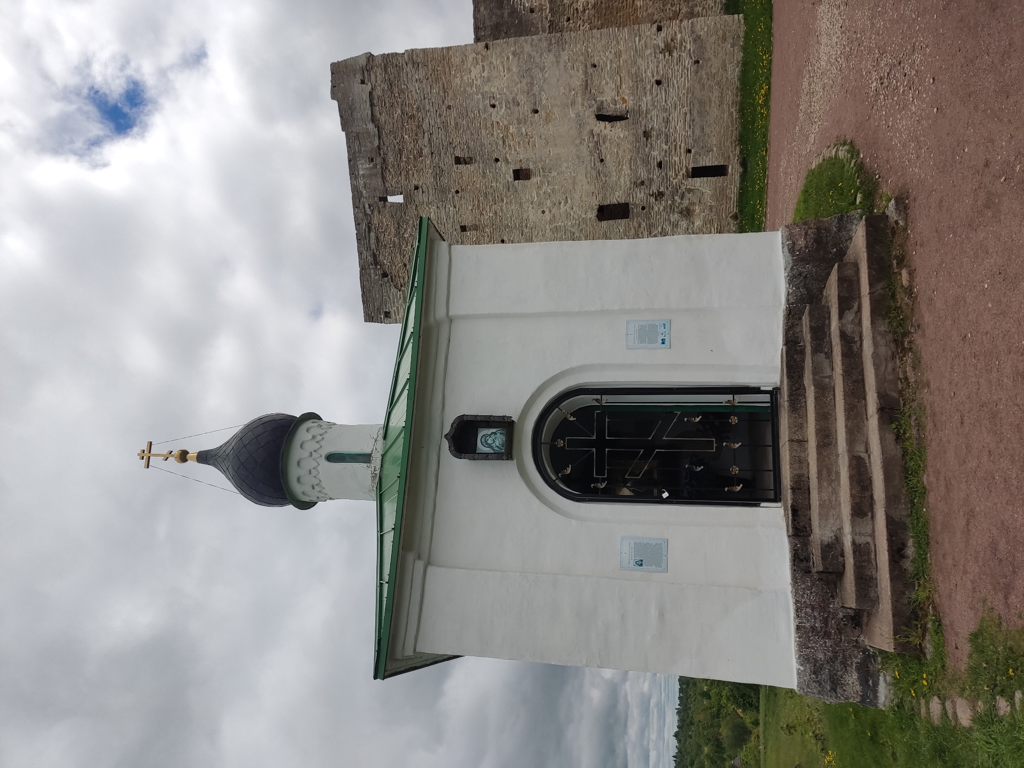 The image depicts a small white Orthodox chapel located in Izborsk, Russia. The chapel has a black dome topped with a gold cross. A stone fortress wall stands adjacent to the chapel. The entrance to the chapel is a dark metal door with a cross design. There are several stone steps leading up to the entrance. The surrounding environment includes a dirt path, grass, and an overcast sky. No people are visible in the image.