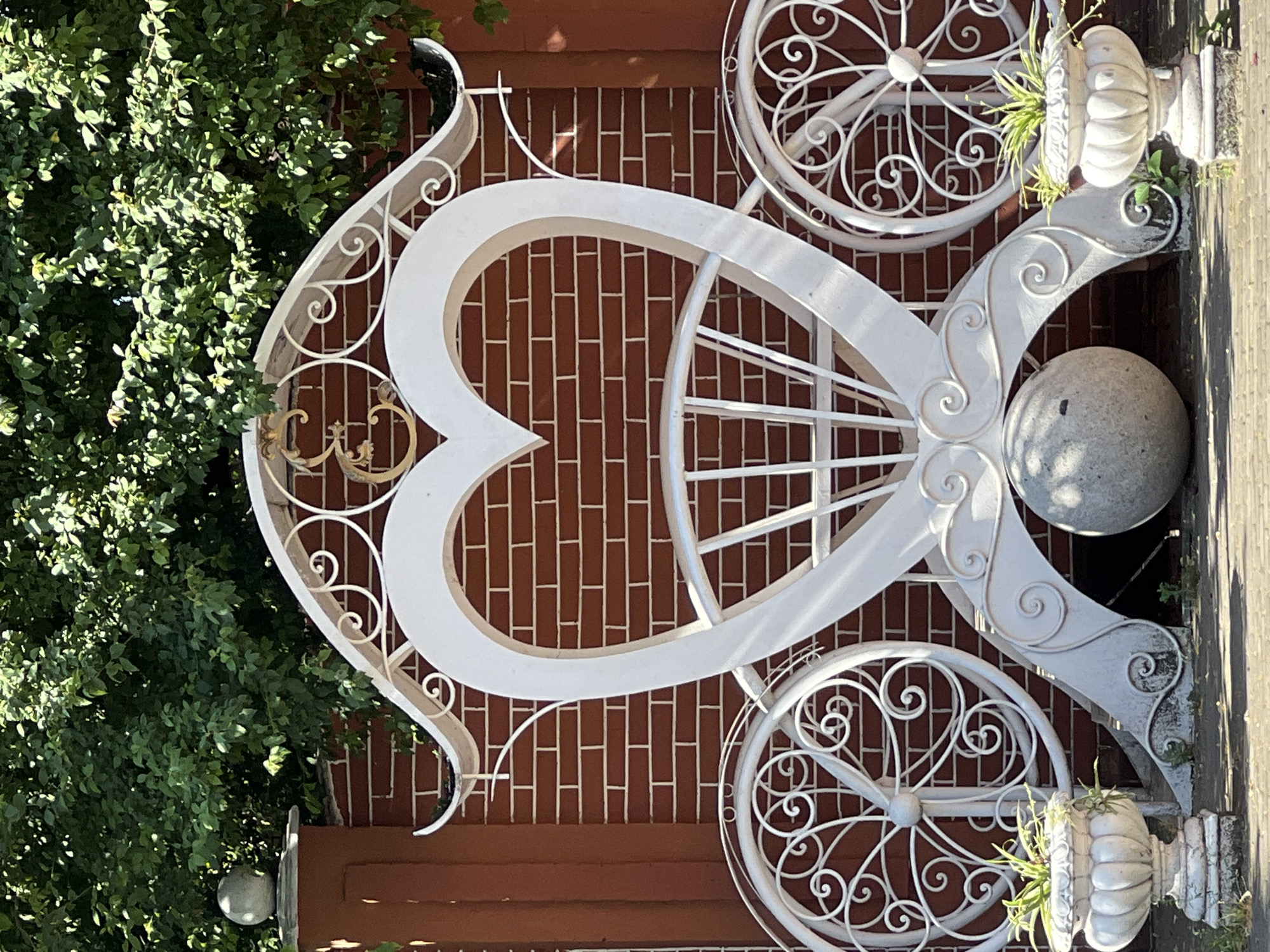 This is a medium shot, eye-level, vertical, outdoor photograph of a white, ornate, wrought-iron decorative structure resembling a carriage or a large heart. The structure is positioned in front of a red brick wall, with lush green foliage visible above and behind it. The carriage-like element has a large heart shape in the center, supported by decorative scrollwork and two large, spoked wheels on either side, also made of wrought iron with intricate swirling patterns. At the base of the structure, there is a large, grey, spherical stone. To the left and right of the main structure, there are white, classical-style planters with green plants. The ground in front appears to be paved with interlocking bricks or stones. The overall impression is of a decorative garden or entrance feature.
The location of this decorative carriage is on the territory of the "Chinari" wedding palace in Krasnodar, Russia.