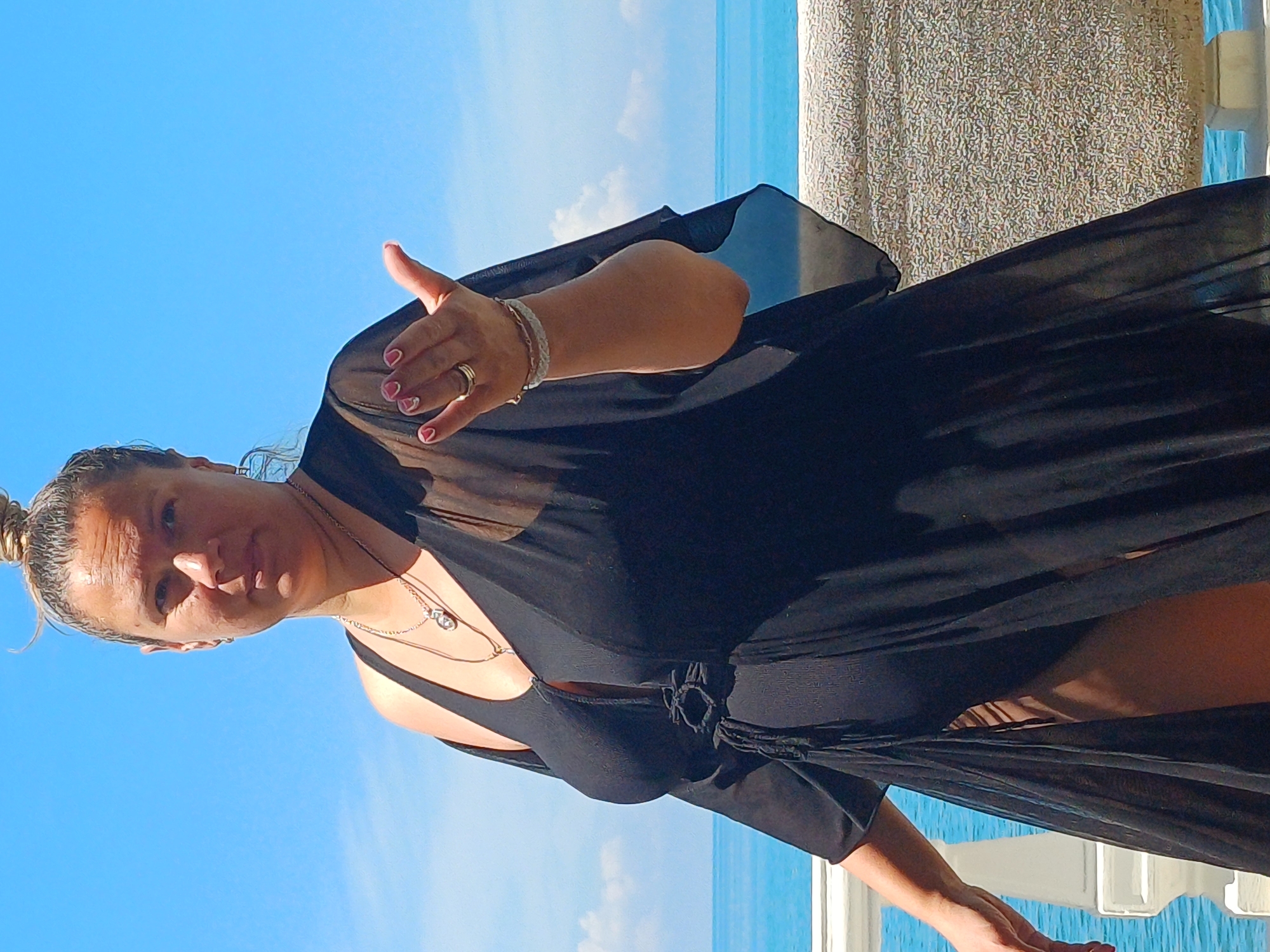 A woman stands on a balcony overlooking the Black Sea in Sochi, Russia. She is wearing a black sheer cover-up over a black swimsuit. Her hair is pulled up in a bun, and she is wearing necklaces and bracelets. The sea is blue and the sky is clear. A white railing is visible in the foreground, and a textured wall is behind the woman.
