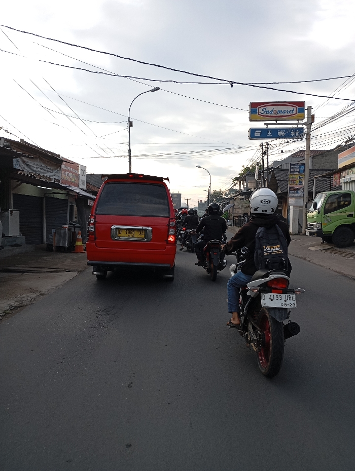 The image shows a street scene in the city of Kutawaringin, Indonesia, located at latitude -6.972 and longitude 107.532. A person wearing a white helmet and a black jacket with a backpack is riding a white motorcycle on a paved street. The motorcycle has a black license plate with the letters "D 4193 JBE" and the numbers "08-20". The person is riding the motorcycle toward the camera. There are other motorcycles in the background. There is a red van parked on the right side of the street. The van has a black license plate with the letters "D 1883 XE". There are buildings on both sides of the street. There is a sign for "Indomaret" on the right side of the street, indicating a local convenience store. The sky is overcast.