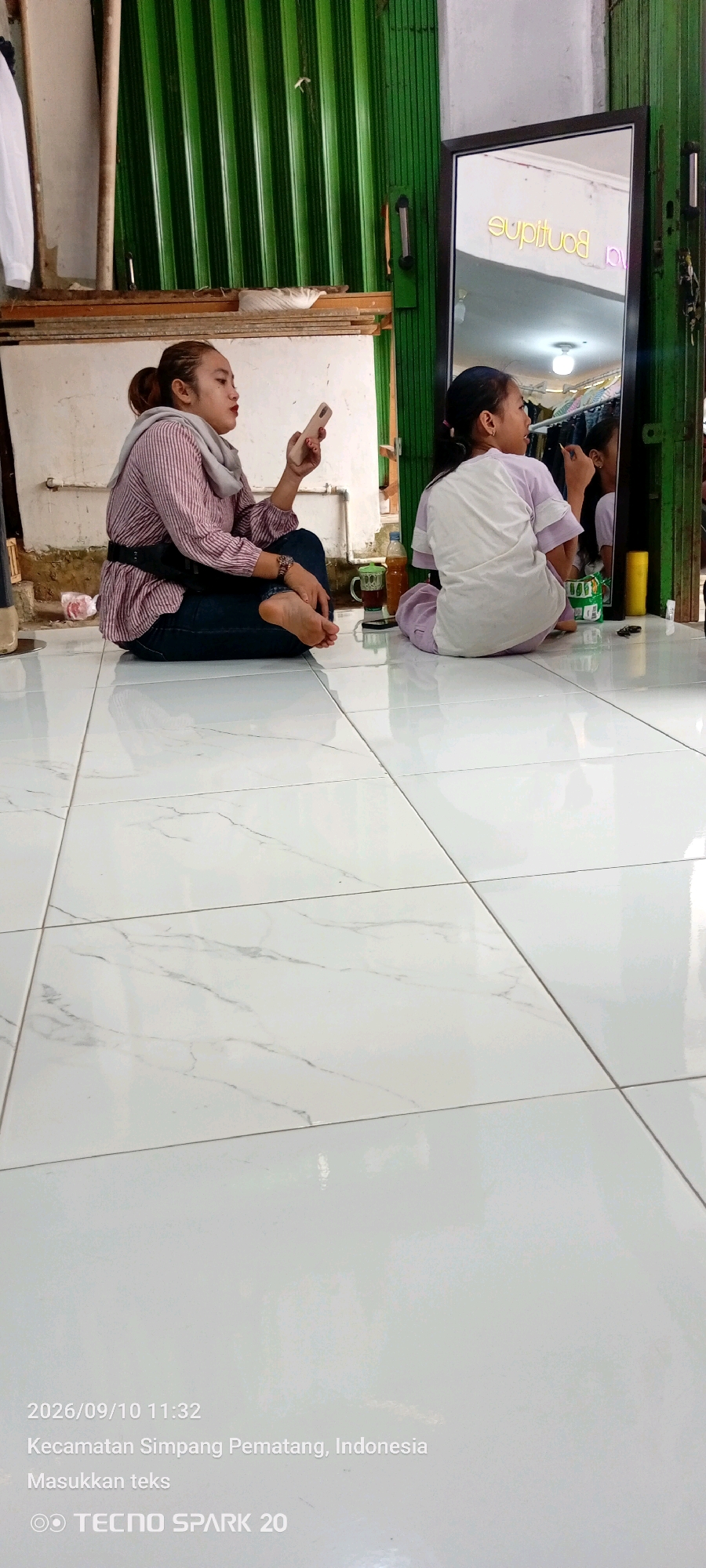 The image depicts an indoor scene with two individuals sitting on a white tiled floor. The individual on the left is a woman with her hair in a bun, wearing a striped shirt, dark pants, and a grey scarf. She is holding a mobile phone in her right hand and looking down at it. To her right, a younger individual, possibly a child or teenager, is sitting on the floor facing a large mirror. This second individual is wearing a light-colored t-shirt and light-colored pants and appears to be applying makeup, looking at their reflection in the mirror.
The environment includes a large, framed mirror reflecting a neon sign that reads "boutique" in yellow lettering. Behind the individuals and the mirror, there is a corrugated green wall, possibly a garage door or siding. To the left of the woman, there is a white board and some wooden planks. On the floor near the individuals, there are a few objects, including a glass with a dark liquid and a bottle.
The timestamp at the bottom left of the image indicates "2026/09/10 11:32" and the location is identified as "Kecamatan Simpang Pematang, Indonesia". The bottom right corner shows "TECNO SPARK 20". The overall scene appears to be casual and domestic or a small retail space, possibly a dressing room or a shop's back area.