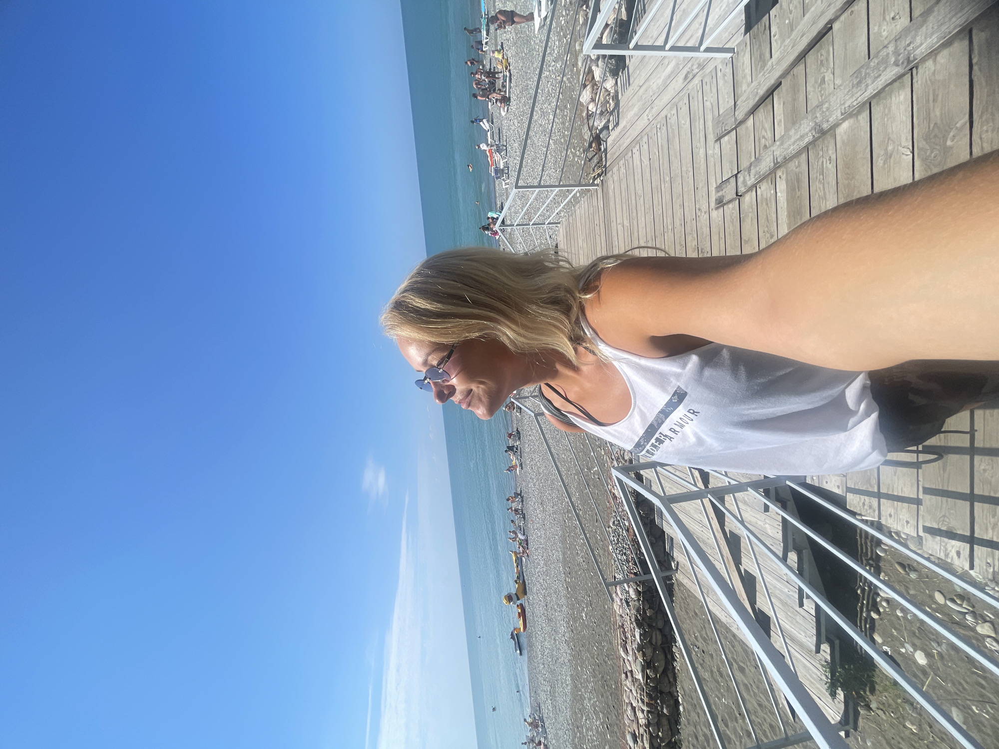 A woman with blonde hair and sunglasses takes a selfie on a wooden boardwalk on a sunny day at the beach. She is wearing a white tank top and her arm is extended holding the camera. The boardwalk has white railings and leads down to a pebble beach. In the background, the sea is calm and blue, with several people sunbathing and some inflatable toys on the shore. The sky is clear and blue. This scene is located in Giachrypsh, Abkhazia.