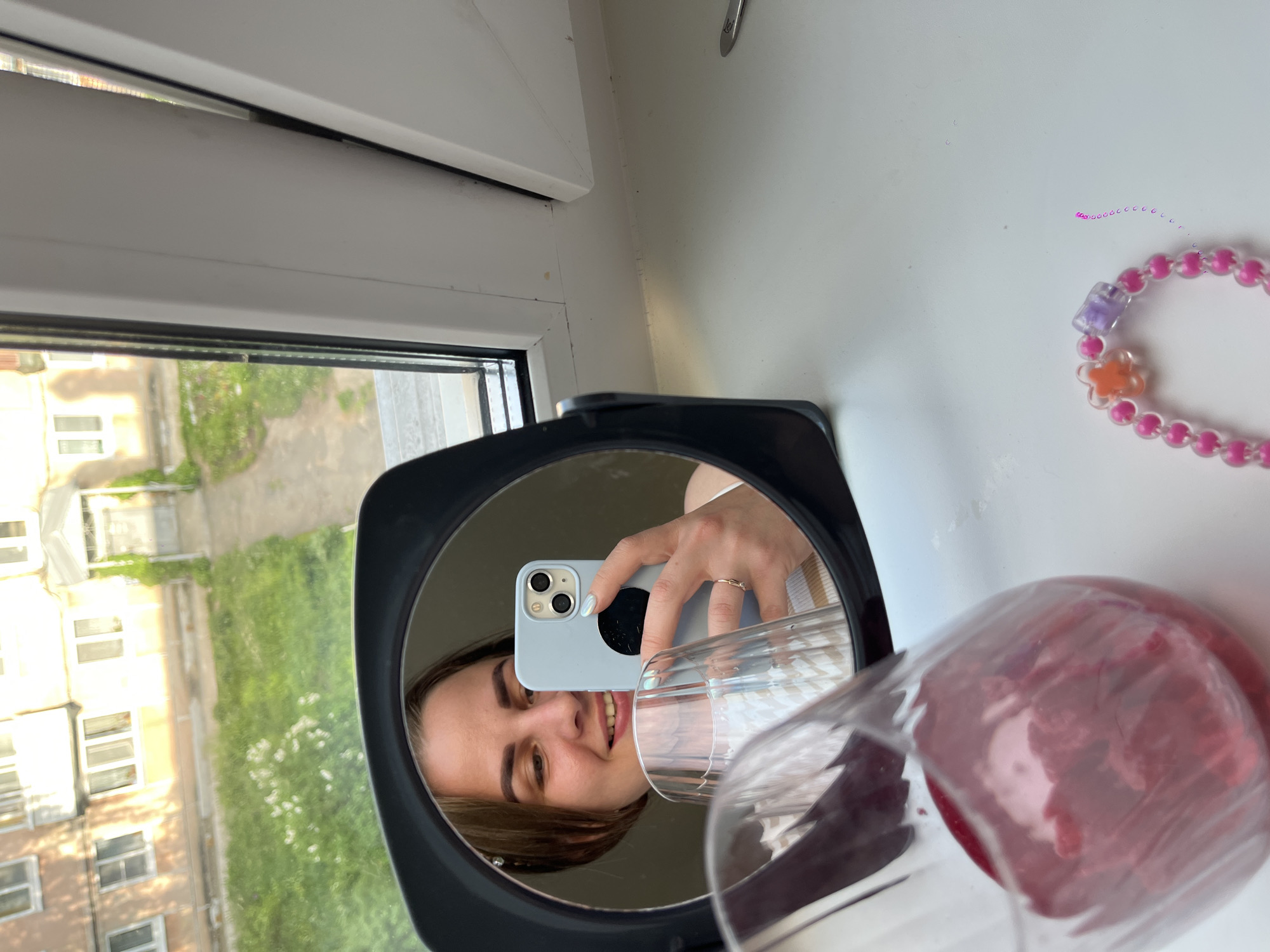 A woman is taking a picture of herself in a mirror. The mirror is black and round and sits on a white surface. In the mirror, she holds a blue phone with three camera lenses. She is smiling. A partially visible glass with a reddish liquid sits beside the mirror. A pink beaded bracelet is on the surface beside the mirror. A window is visible in the background reflecting the cityscape of Izhevsk, Russia. Buildings and greenery can be seen through the window. The overall setting appears to be indoors, possibly a windowsill.