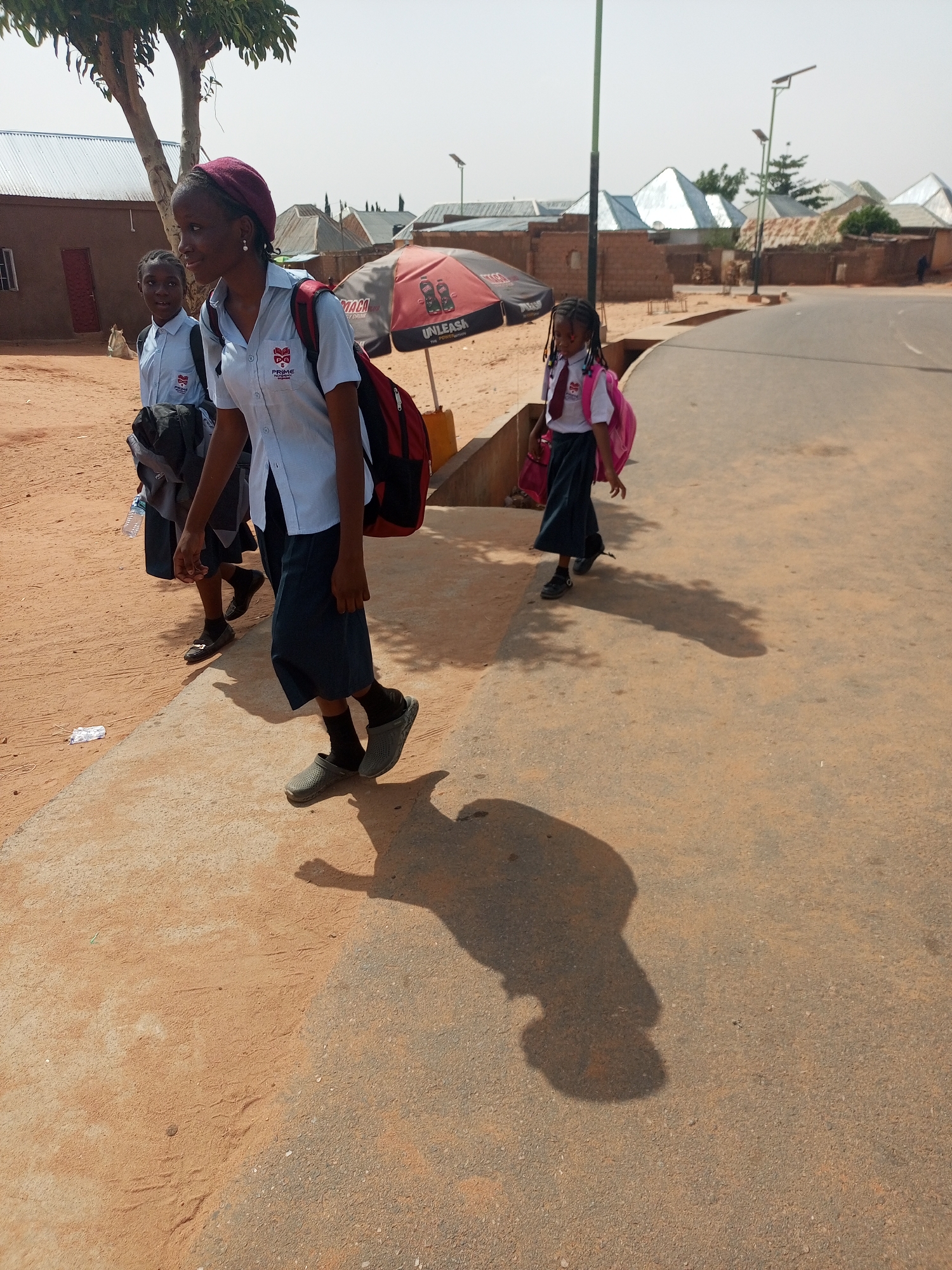 Three girls in school uniforms walk along a paved road in Gombe, Nigeria. The girls are wearing light blue shirts with a school logo, dark skirts, and backpacks. A street lamp is in the background and various buildings in the background have corrugated metal roofs. The scene is bathed in bright sunlight.