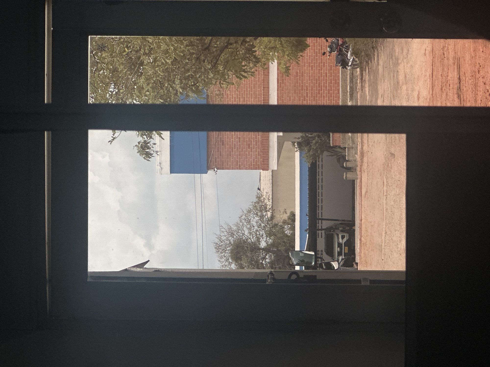 This media file shows a view through a window or door frame, with horizontal bars dividing the scene. The image appears to be taken outdoors, possibly in a rural or semi-rural area given the presence of dirt pathways and buildings with exposed brick.
In the upper portion of the image, a tree with green foliage is visible, along with a brick building and a motorcycle parked on a dirt path. The sky is partly cloudy. The lower portion of the image shows more of the brick building, a blue-colored section, and what looks like a large truck or utility vehicle with visible equipment. A bare tree with thin branches is also present. The overall scene suggests daytime. There is no discernible text in the image. The perspective suggests the viewer is looking out from a shaded interior. The scene is consistent with a location in Juan Mina, Colombia, characterized by its climate and typical structures.