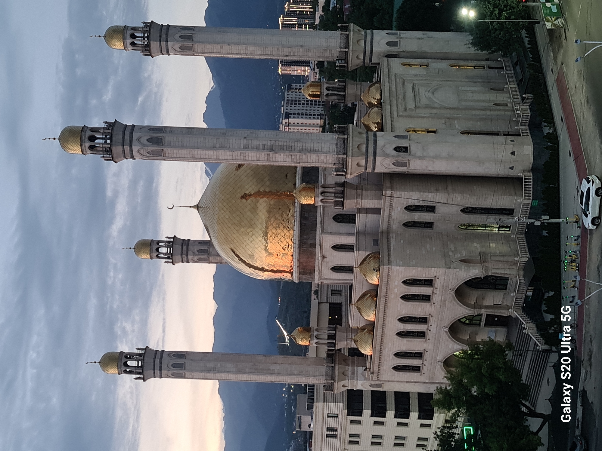This image captures the impressive architecture of the Central Mosque of Almaty, Kazakhstan, during what appears to be dusk or dawn. The mosque is a prominent structure with a large, ornate golden dome and four tall minarets, also topped with golden domes. The building itself is constructed from light-colored stone and features arched windows and intricate detailing.
In the background, a range of imposing mountains can be seen, their peaks silhouetted against a cloudy sky that displays soft hues of orange and purple, indicative of either sunrise or sunset. The city of Almaty sprawls in the middle ground, with modern buildings, some illuminated by lights, interspersed with trees and greenery.
In the foreground, a white car is parked on a paved road, and a red pedestrian path runs along the edge of the mosque's property. Several electric scooters are also visible, lined up on the sidewalk. The scene is devoid of any people actively engaged in the image, suggesting a quiet moment in the city. The lighting suggests the image was taken from an elevated position, likely a building overlooking the mosque and its surroundings. The bottom left corner of the image includes a watermark indicating "Galaxy S20 Ultra 5G".
