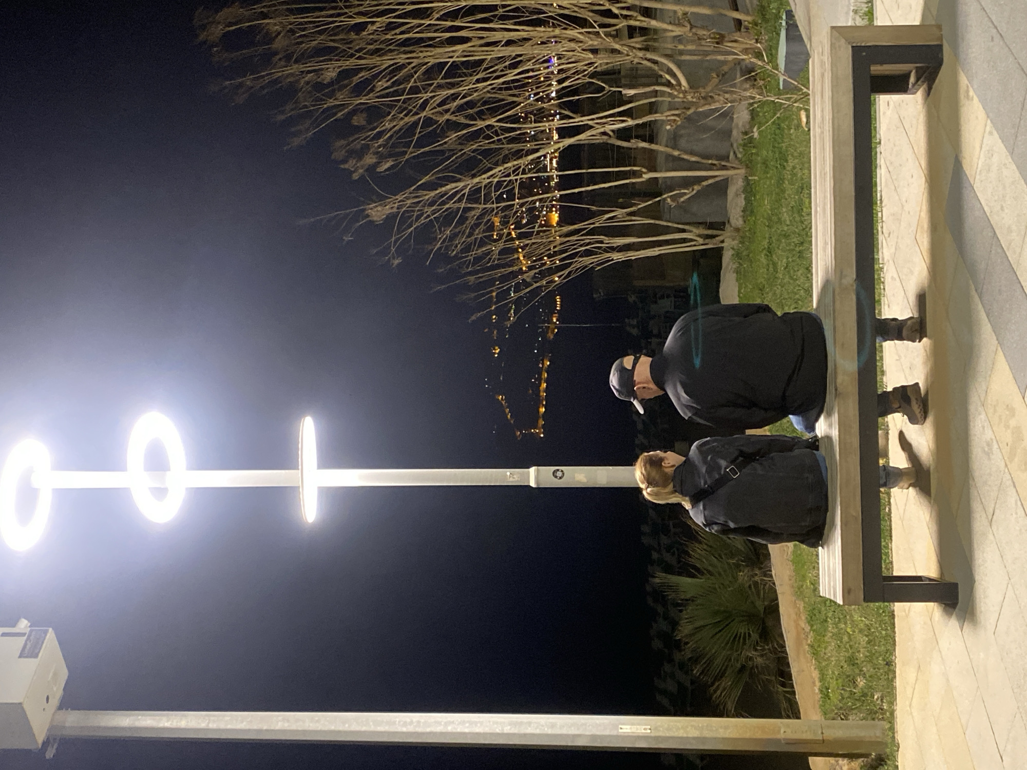 A nighttime scene in Alanya, Turkey, features two people sitting on a bench next to a modern lighting fixture. The light fixture is a tall pole with three circular LED lights on top and a horizontal bar extending from the middle. A man wearing a black cap and jacket sits to the right, while a woman in a dark jacket is seated beside him. Behind them, a tree with bare branches is visible, with blurred city lights in the background. The bench is constructed of light-colored wood with dark metal supports, situated on a tiled walkway bordering a patch of grass.