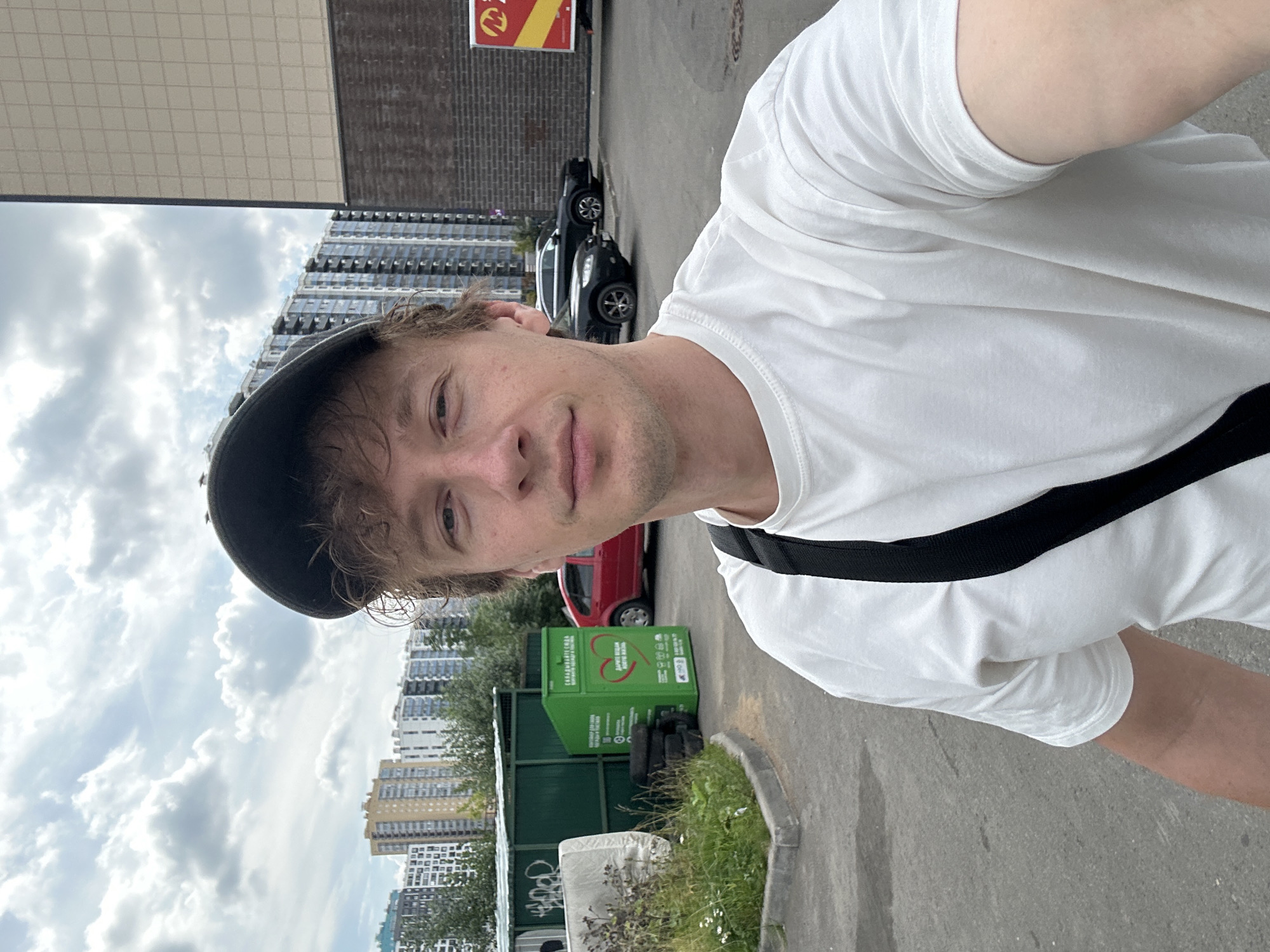 A man wearing a white t-shirt and a dark baseball cap takes a selfie outdoors. He has light brown, curly hair and a light complexion. A black strap is visible across his chest. In the background, there are several apartment buildings and a red car. To the left, there is a green recycling bin with a heart symbol and Cyrillic text. The ground is paved and there is some greenery to the left of the recycling bin. The sky is cloudy. The location appears to be in Murino, Russia, given the architectural style of the buildings.