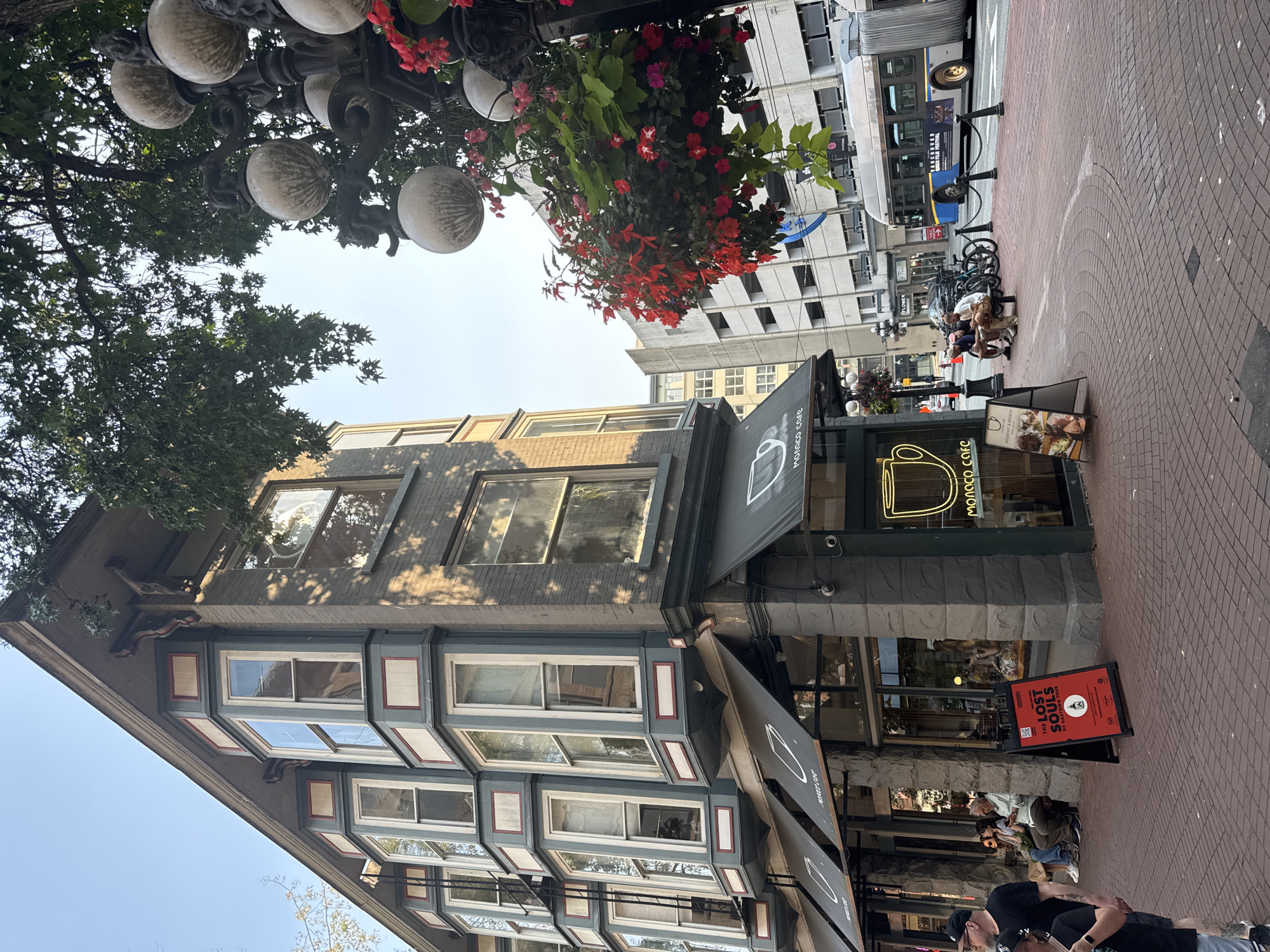 The image depicts a street scene in Gastown, Vancouver, Canada. A historic building with bay windows and a cafe called "Monaco Cafe" occupies the left side of the frame. A street lamp with decorative globes and hanging red flowers stands in front of the building. On the right, a bus with the advertisement "Vancouver Queer Film Festival" is visible on the street. Several people are seated on a bench in front of the cafe, and others are walking on the brick-paved sidewalk. A red A-frame sign for "The Lost Souls of Gastown Tour" is propped up on the sidewalk in front of the cafe. The sky is clear and blue, and the sun casts shadows on the building.