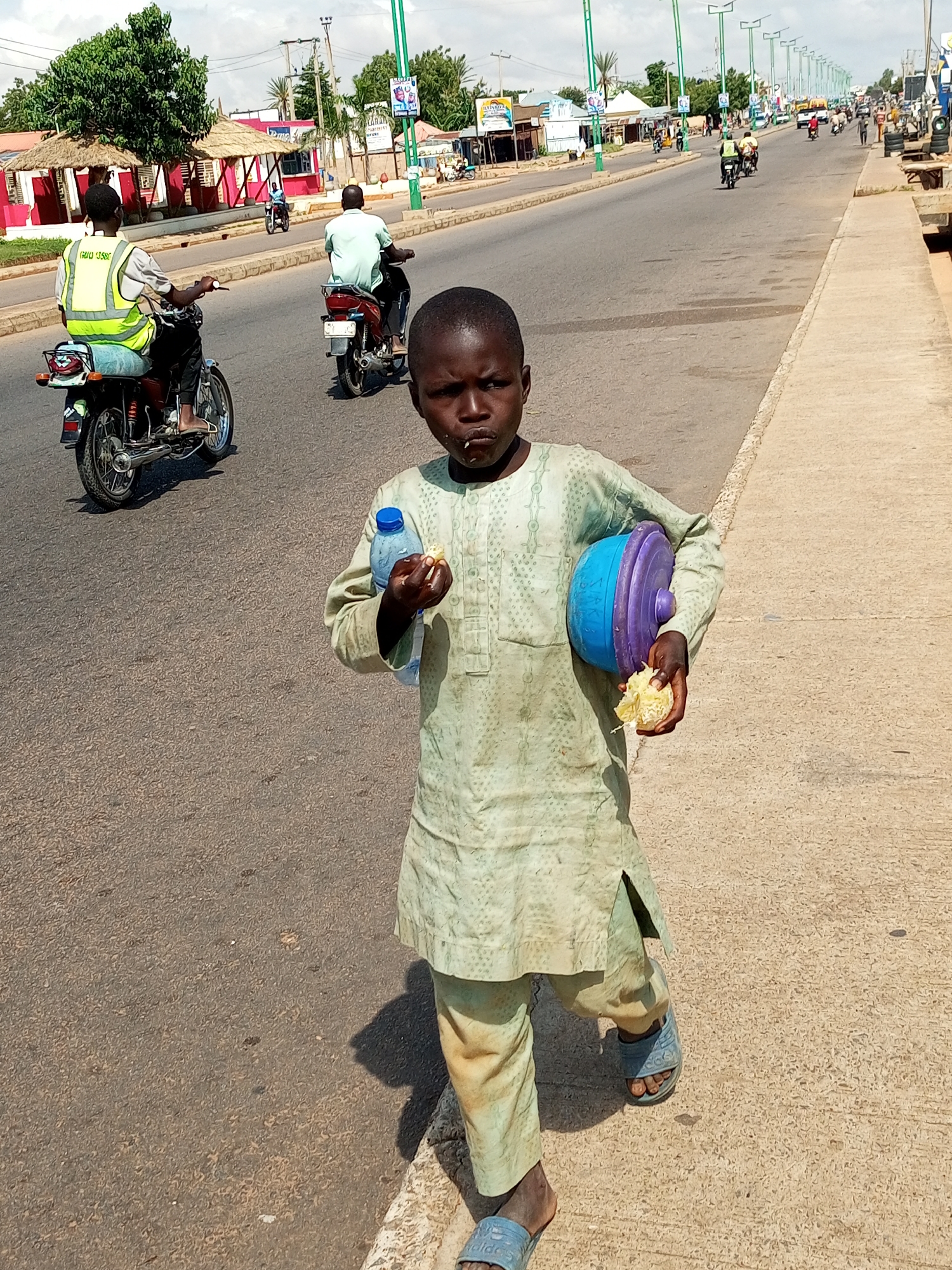 A young boy, appearing to be approximately 8-10 years old, walks along a paved road in Tempure, Nigeria. He is wearing a pale green traditional tunic and trousers, and blue flip-flops. In his right hand, he holds a piece of food, and in his left hand, a blue plastic container with a purple lid, as well as a partially consumed bottle of water. He is looking towards the camera. The road is a busy thoroughfare with several motorcycles and vehicles. In the background, there are buildings with thatched roofs and modern structures, suggesting a mix of traditional and contemporary architecture. Streetlights line the road. The weather appears to be clear and sunny.