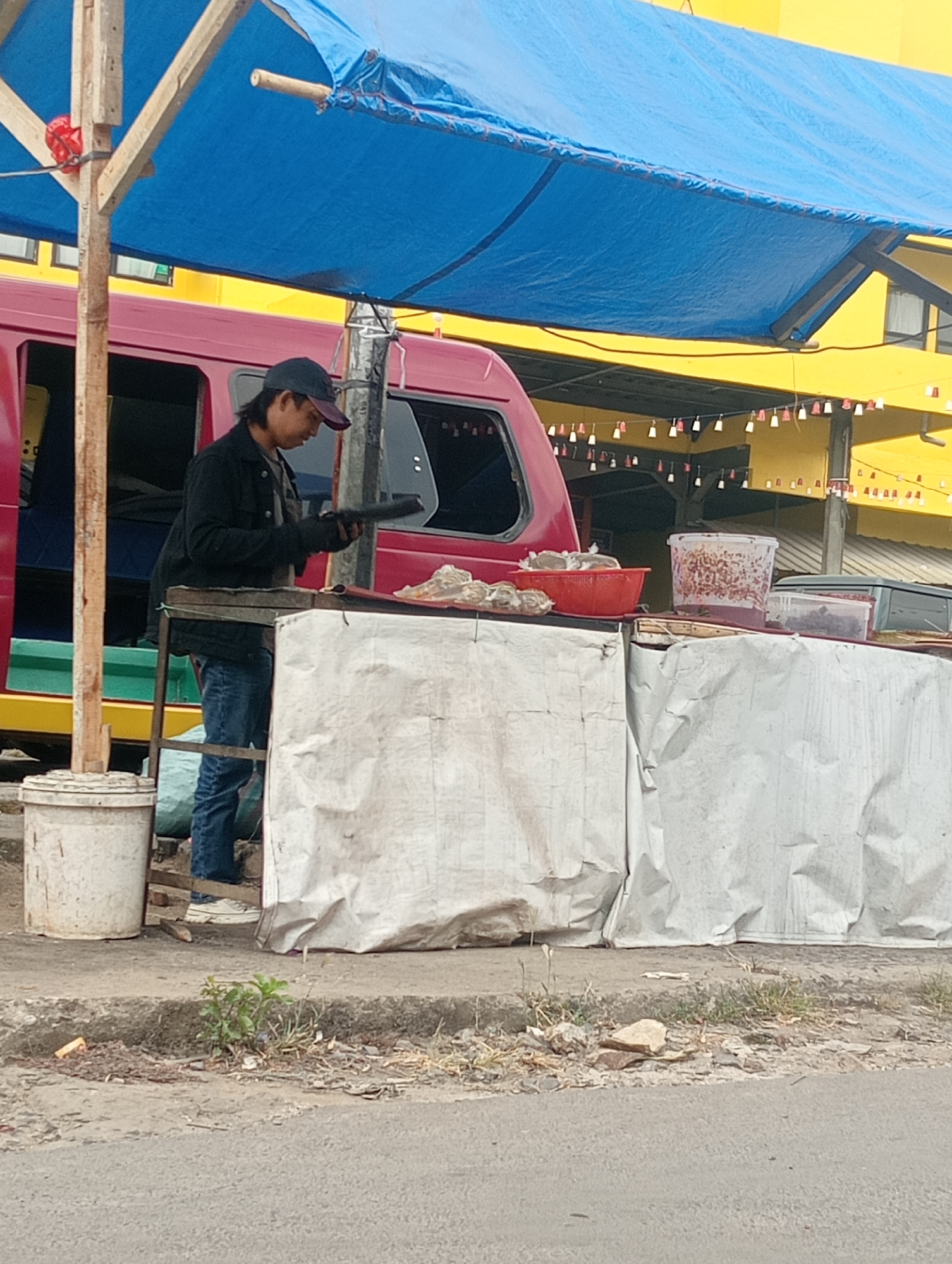 A person wearing a dark jacket, a dark baseball cap, and blue jeans stands behind a makeshift stall on a street in Bandar Lampung, Indonesia. The stall has a blue tarp overhead, supported by wooden poles, and its front is covered with a wrinkled, off-white material. The person is holding a dark, flat object, possibly a tool or a serving utensil, in their gloved hands. In front of the stall are various containers and a red basket holding small food items. To the left of the stall is a white bucket. The background shows the side of a red vehicle, a yellow building with windows, and strings of small, decorative flags. The ground in front of the stall is paved, with a strip of unpaved earth containing sparse vegetation and debris along the curb. The scene appears to be an outdoor food vending area.