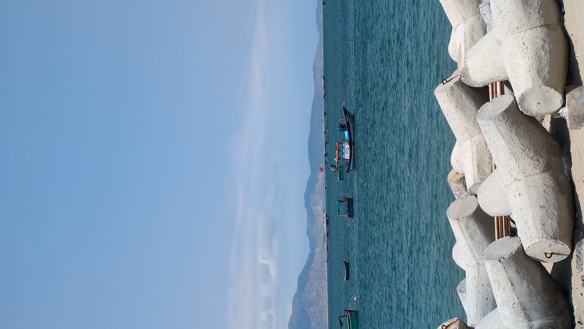 The image shows a seascape in Phan Rang – Tháp Chàm, Vietnam, with concrete tetrapods forming a breakwater on the right side. Boats are floating in the water. Mountains are visible in the distance, and the sky is clear and blue.
