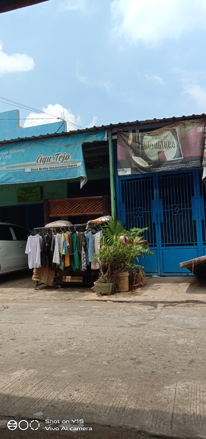 A street scene captures a building adorned with blue paint and banners advertising "Ayu Tejo" and "Nusantara". Laundry hangs from a clothes rack next to potted plants, positioned in front of a blue metal gate. The street is concrete, and the sky is partly cloudy. Text at the bottom of the image indicates the photo was taken with a Vivo AI camera.