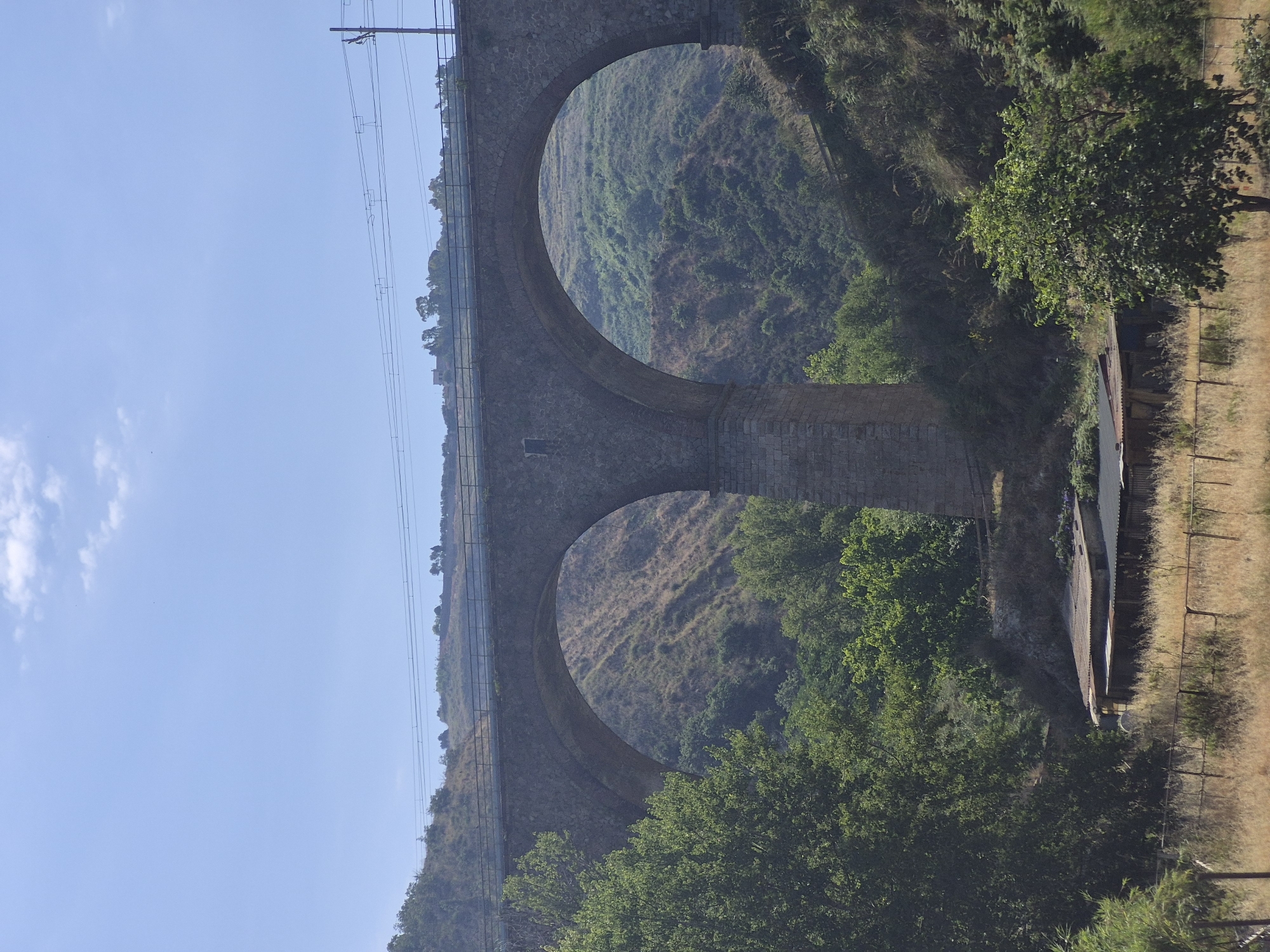 A wide-angle, eye-level, outdoor shot depicts a stone viaduct with two large arches, characteristic of the terrain surrounding Tropea, Italy. The viaduct spans a valley covered in dry brush and green foliage, with trees in the foreground. Overhead power lines run across the top of the viaduct. The sky is clear blue with a few faint clouds. In the lower portion of the image, there are some small, rustic buildings with corrugated metal roofs, surrounded by dry grass and a fence. There are no people visible in the image.