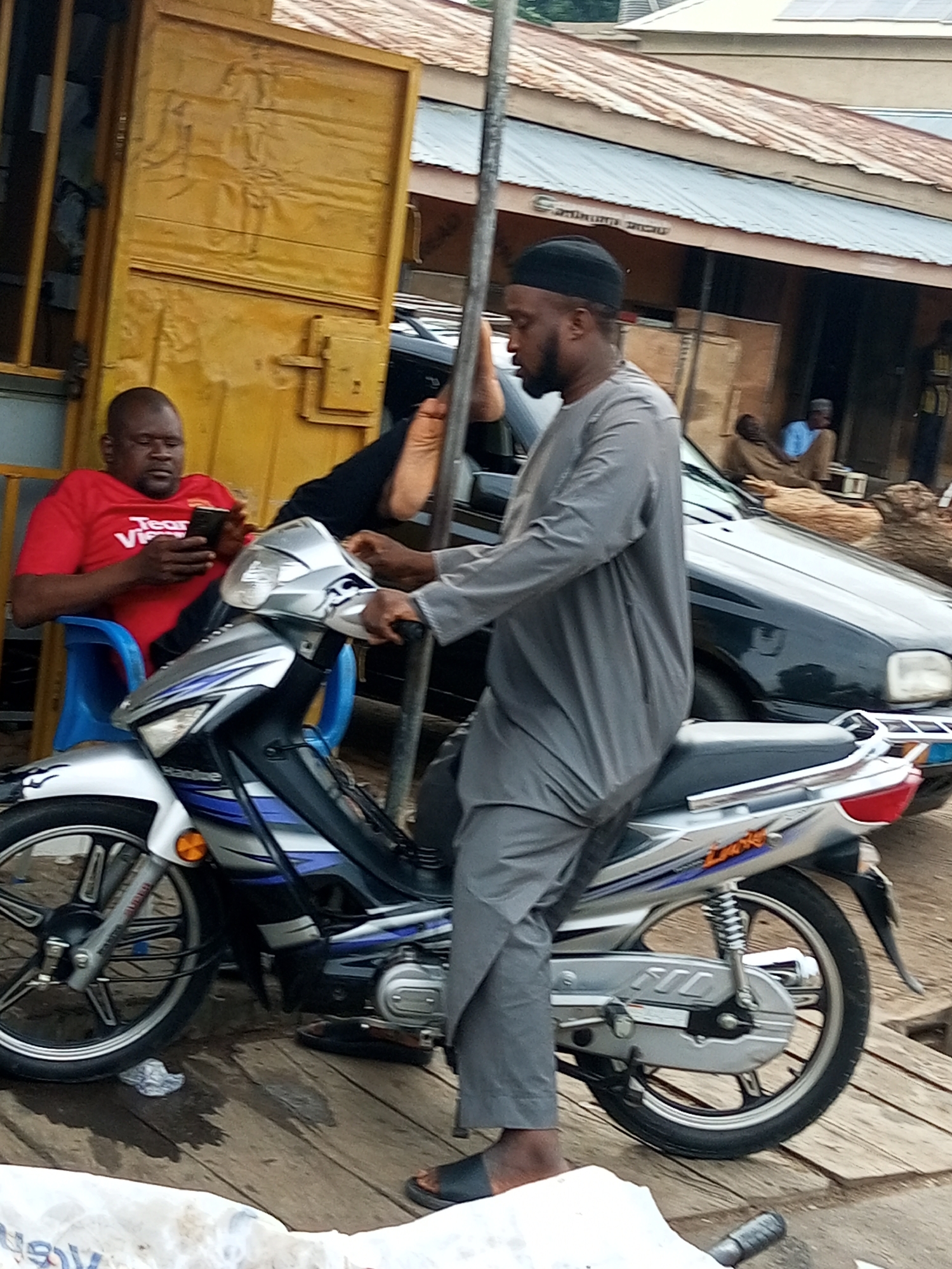 In Arawa, Nigeria, a man wearing a grey thobe and black skullcap sits on a silver and blue motorcycle. The motorcycle is positioned on a wooden surface that appears to be a street or walkway. To the left of the motorcycle, another man in a red t-shirt is seated on a blue plastic chair, looking at his mobile phone. Behind the seated man, a yellow metal structure, possibly a shop or doorway, is visible. Further in the background, a black car is parked, and other structures with corrugated metal roofs suggest an urban environment. The overall scene depicts a street-level view with people and vehicles typical of a Nigerian city.