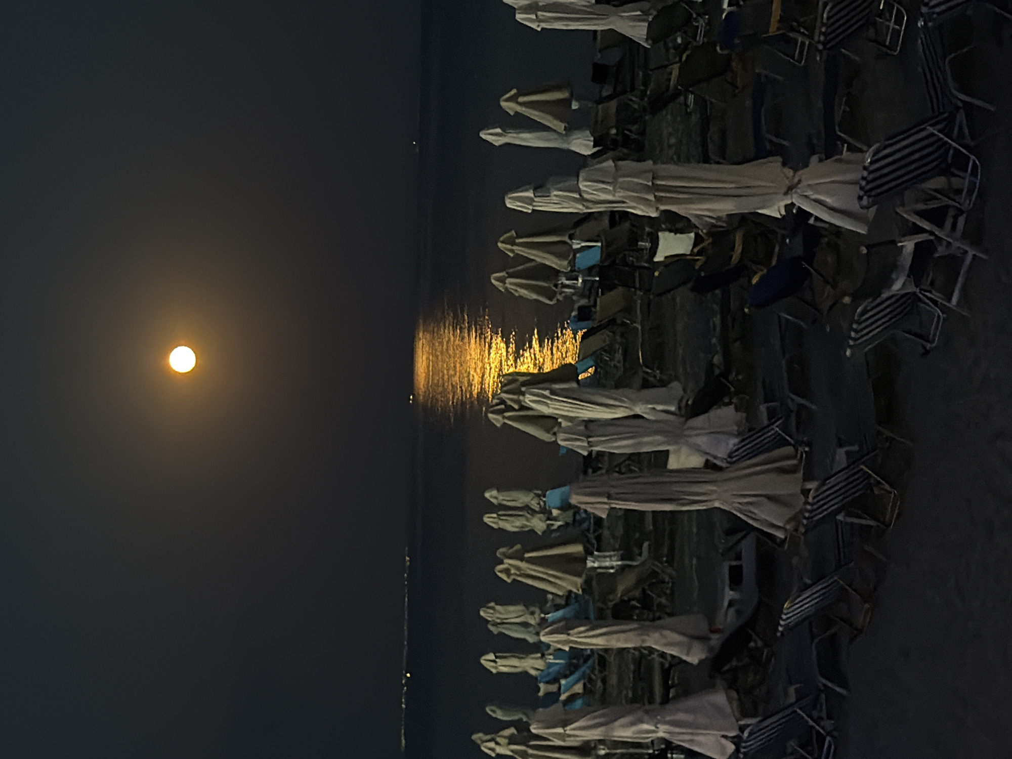 The image depicts a serene night scene at a beach in Nea Kallikratia, Greece, illuminated by a bright, full moon. The moon hangs in the dark sky, casting a warm, golden glow onto the calm sea, creating a shimmering reflection. In the foreground, numerous beach chairs and umbrellas are neatly arranged on the sand, most of them closed and covered, indicating the beach is not actively in use at this hour. The chairs are of various designs, with some featuring striped patterns. The overall atmosphere is peaceful and tranquil, highlighting the natural beauty of the coastal landscape under the moonlight.