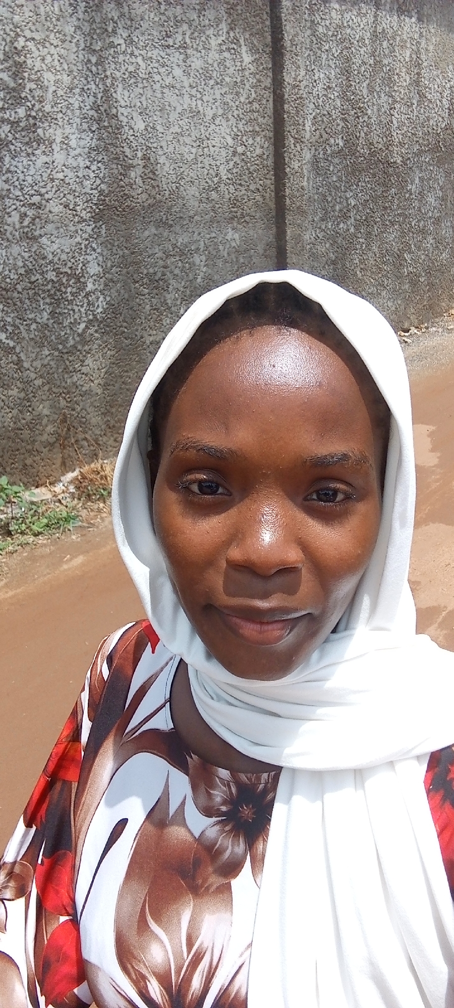 A woman is centered in the frame, wearing a white headscarf and a floral print shirt. The background reveals a textured gray wall and a reddish-brown dirt road, suggesting an outdoor location. Given the prompt's request for a location in Abuja, Nigeria, it is plausible that the scene is set on a street in a residential or suburban part of the city with unpaved roads and simple building materials. There are no other people visible in the shot, nor are there clear interactions. The woman's expression is neutral, without an obvious emotional display.